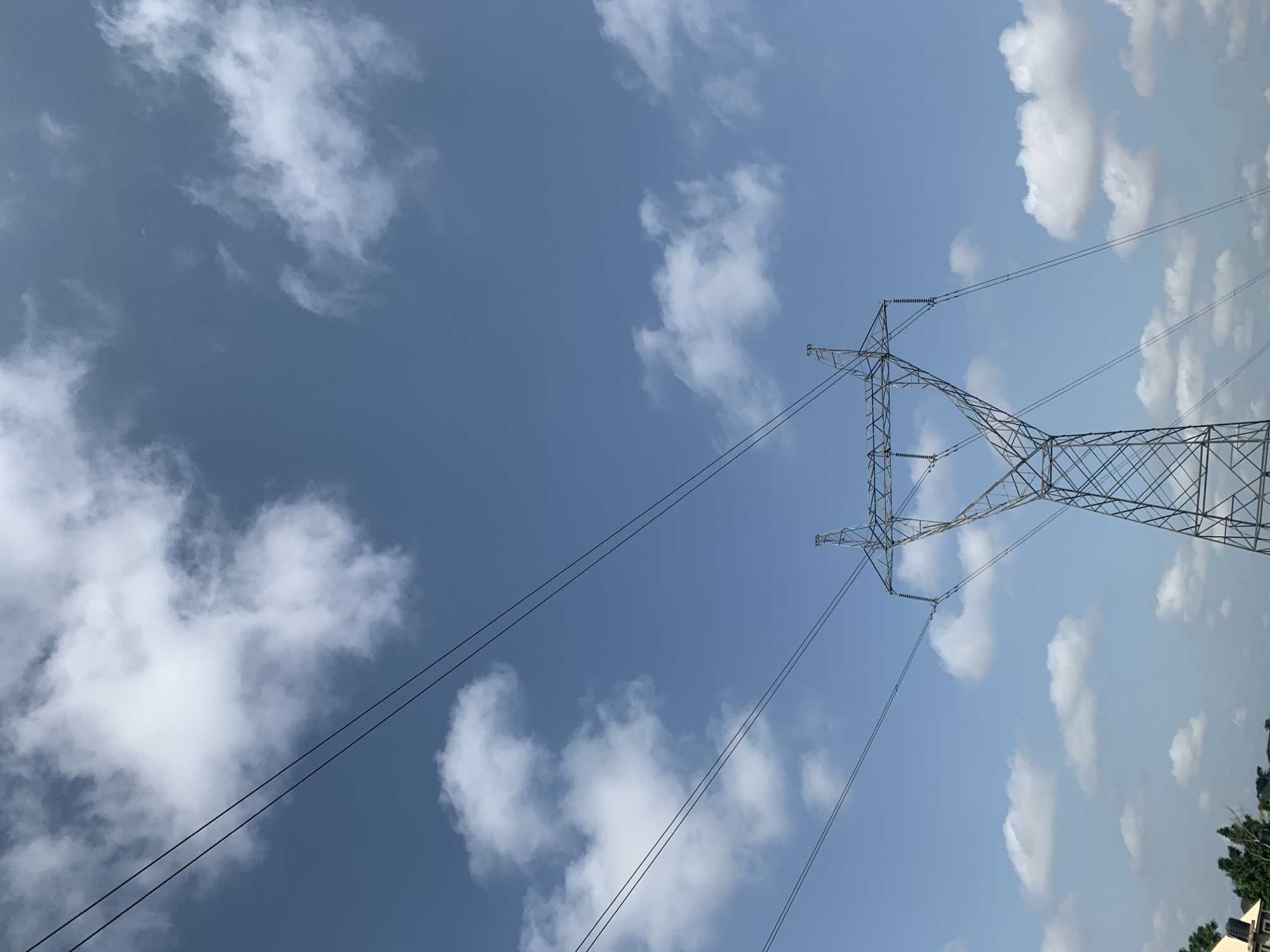 A close-up, low-angle shot captures a large, lattice-style metal power tower against a bright blue sky dotted with scattered white clouds. Several thick, black electrical wires extend diagonally across the frame from the top left, connected to the power tower. The lower portion of the image shows the base of the tower, which is a complex network of intersecting metal beams. In the far lower left corner, a sliver of a residential area is visible, with the roof and part of a building, along with green trees. The overall scene suggests an outdoor, possibly semi-urban or rural environment on a clear day.