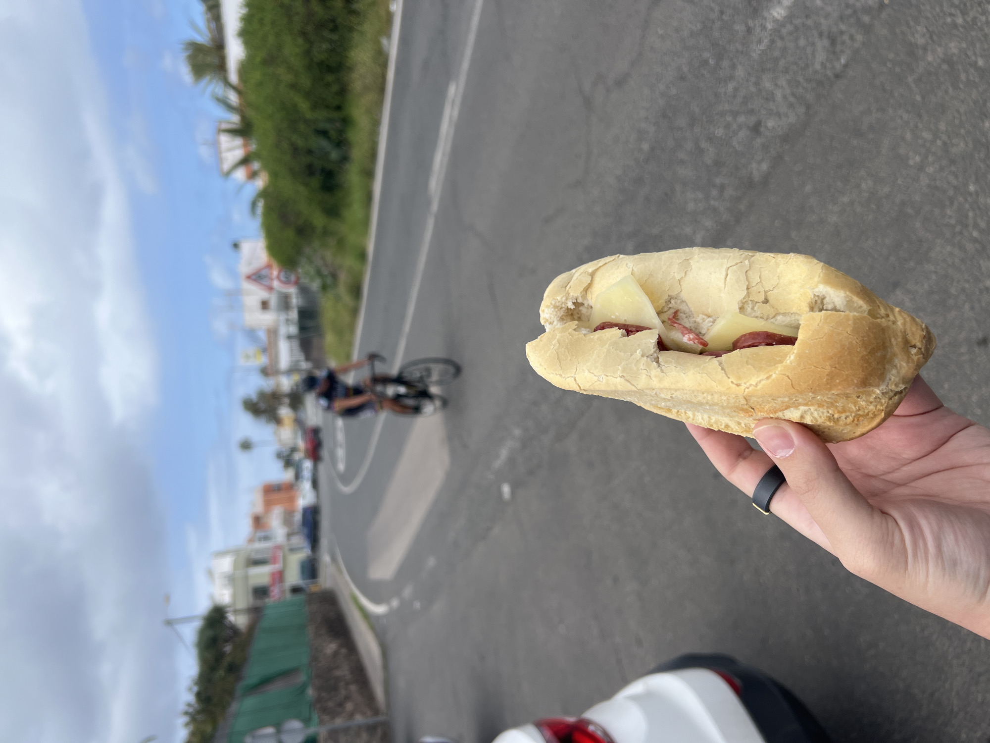 A person is holding a sandwich with cheese and sausage in their hand while standing on the side of a road in La Aldea de San Nicolás, Spain. In the background, a cyclist is riding on the road, and buildings and greenery are visible. The sky is partly cloudy. The road is paved and has lane markings. A white and red vehicle is partially visible in the lower part of the image. The overall setting suggests an outdoor scene during daylight hours.