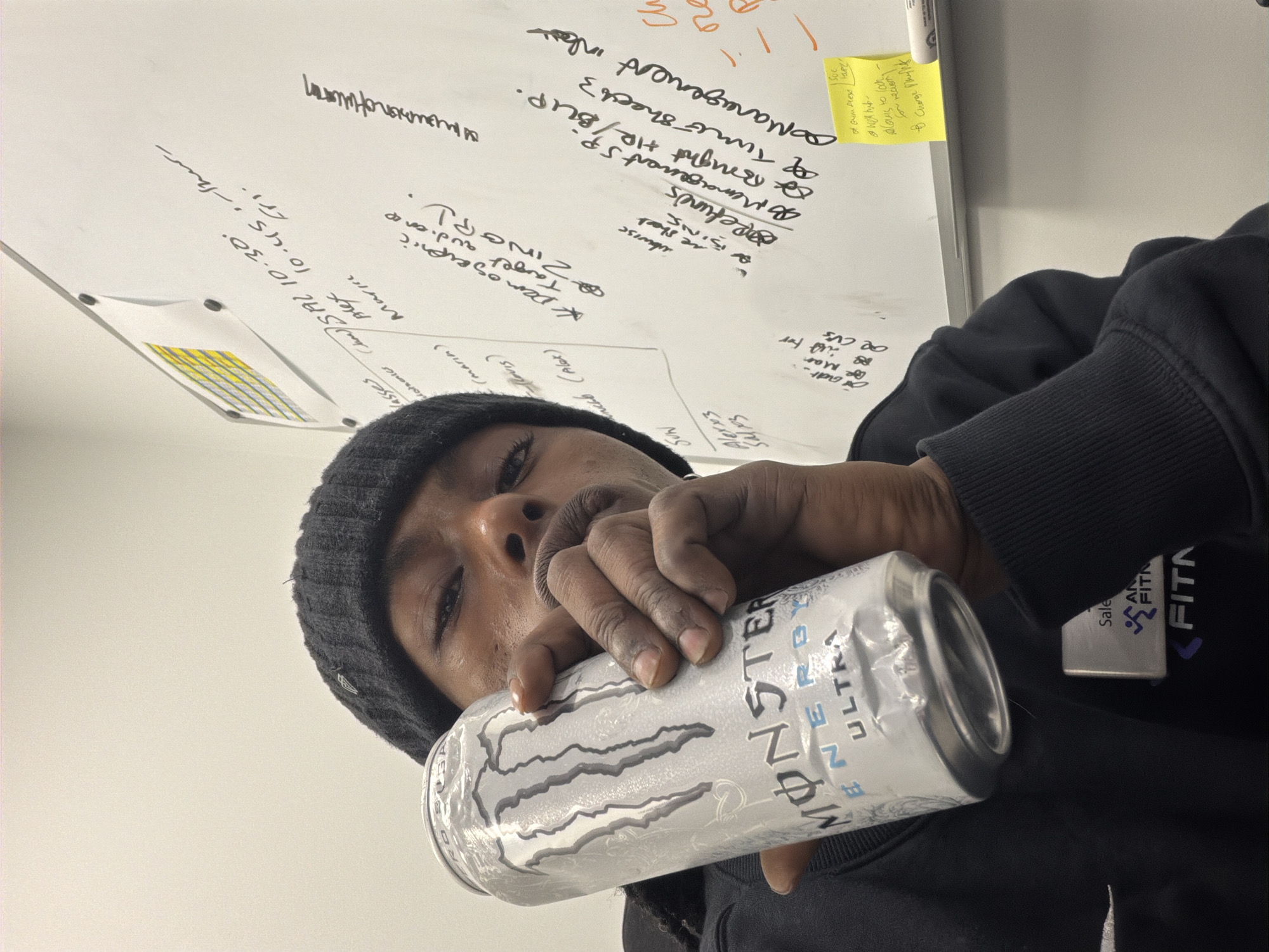 A person wearing a black beanie and black sweatshirt is holding a can of Monster Energy Ultra drink in their hand. The person's face is partially visible, and they appear to be indoors. Behind them is a whiteboard with handwritten notes and a yellow sticky note. A company logo, "AN FITN," is visible on the sweatshirt. The location is not discernible from the image.