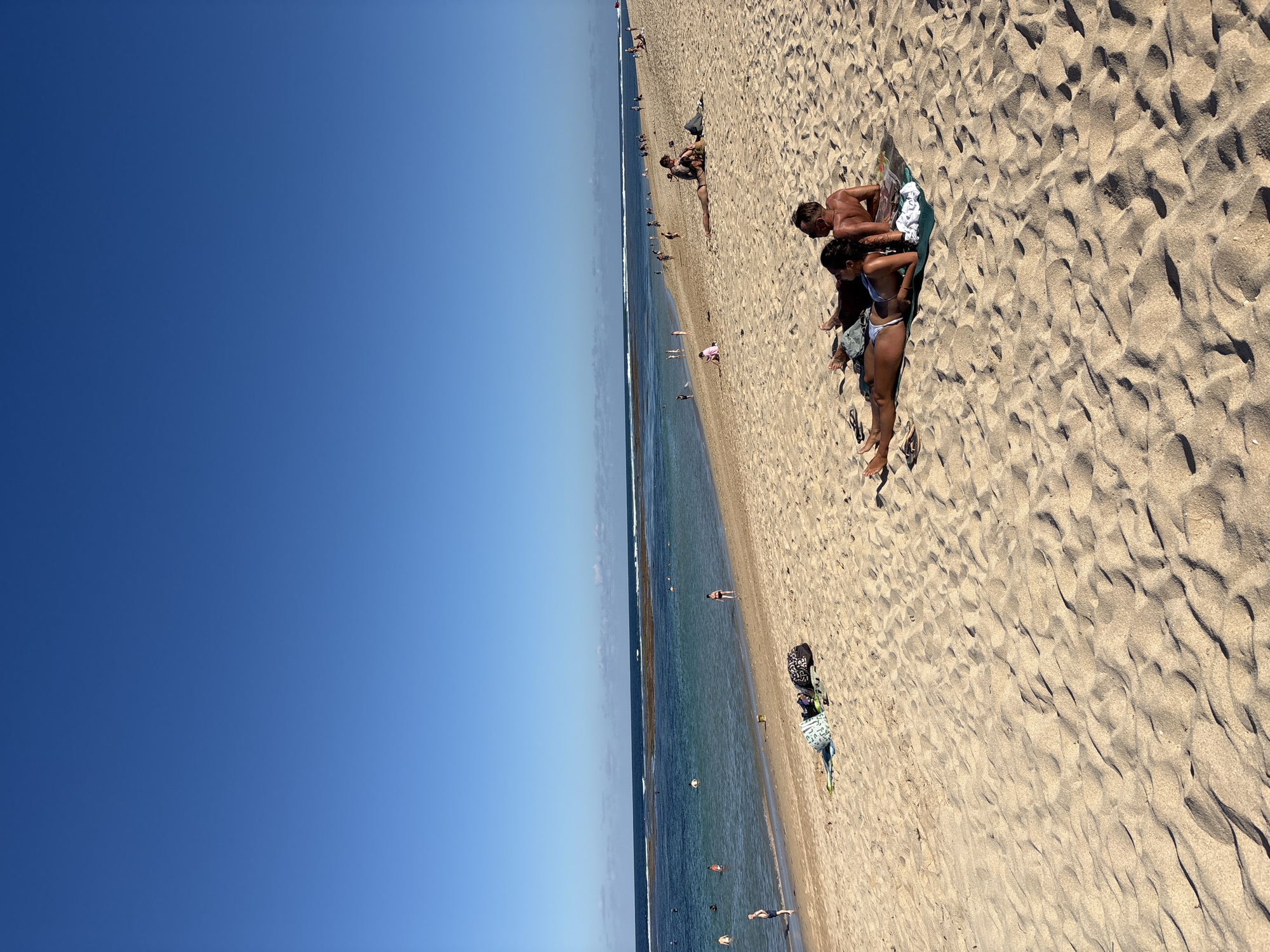 The image depicts a beach scene in Kuta Selatan, Indonesia. The sky is clear and blue, contrasting with the tan-colored sand. The ocean is visible as a strip between the sky and the beach. People are scattered along the beach, some sunbathing and others swimming in the water. A couple is lying on a towel on the sand. Beach accessories, such as bags and flip-flops, are near them.