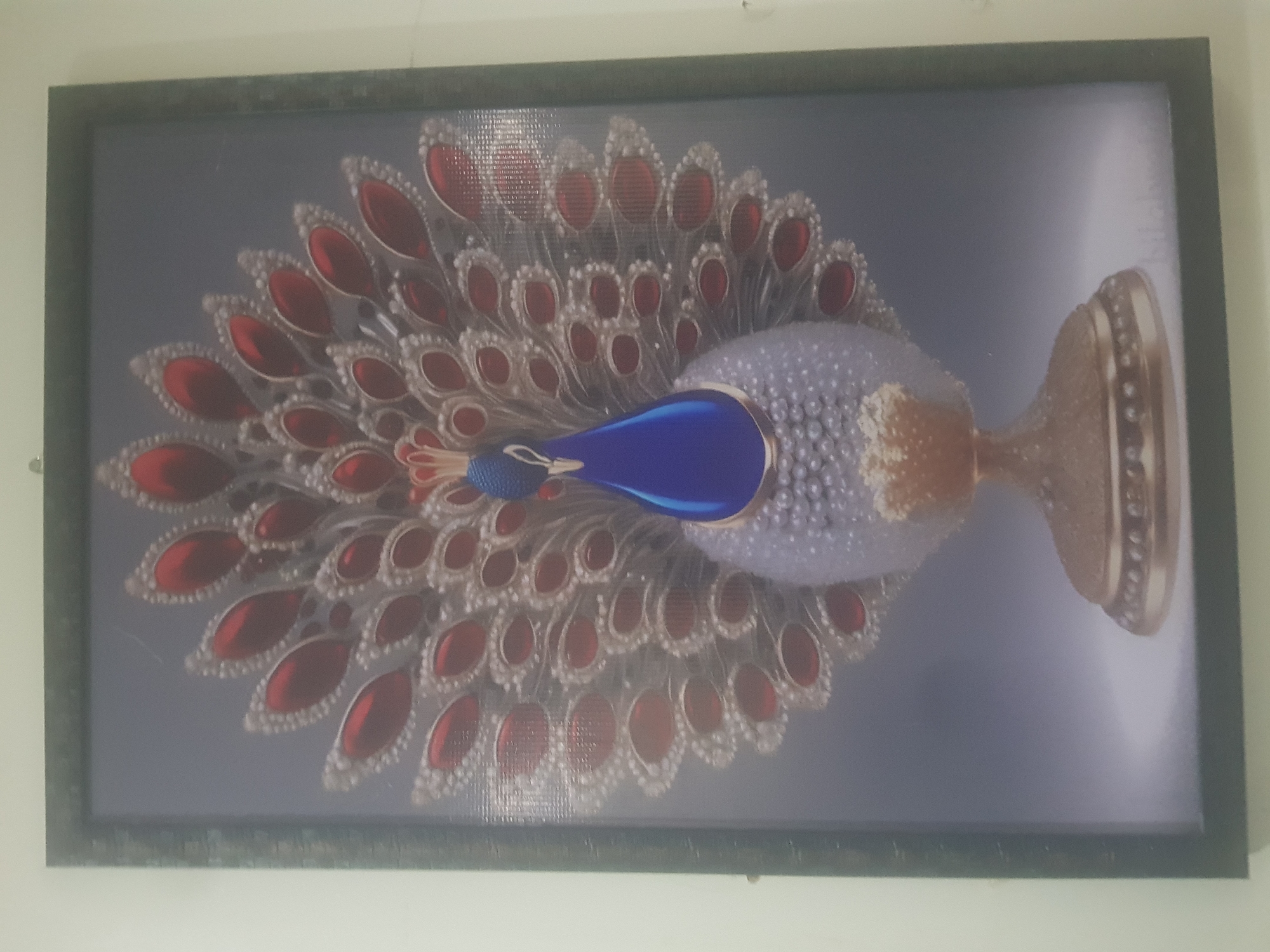 A framed picture of a peacock adorns a wall. The peacock is depicted with its tail feathers spread in a fan-like display. The feathers are adorned with red jewel-like shapes surrounded by intricate silver or white detailing. The body of the peacock is a pearlescent white, with a textured pattern that resembles small beads or pearls. A golden base supports the peacock, which is a pedestal with decorative elements. The background of the image is a gradient of grey, with a soft light illuminating the peacock from the front. The frame of the picture is dark and appears to be made of wood or a similar material. The overall impression is one of opulent and decorative art.