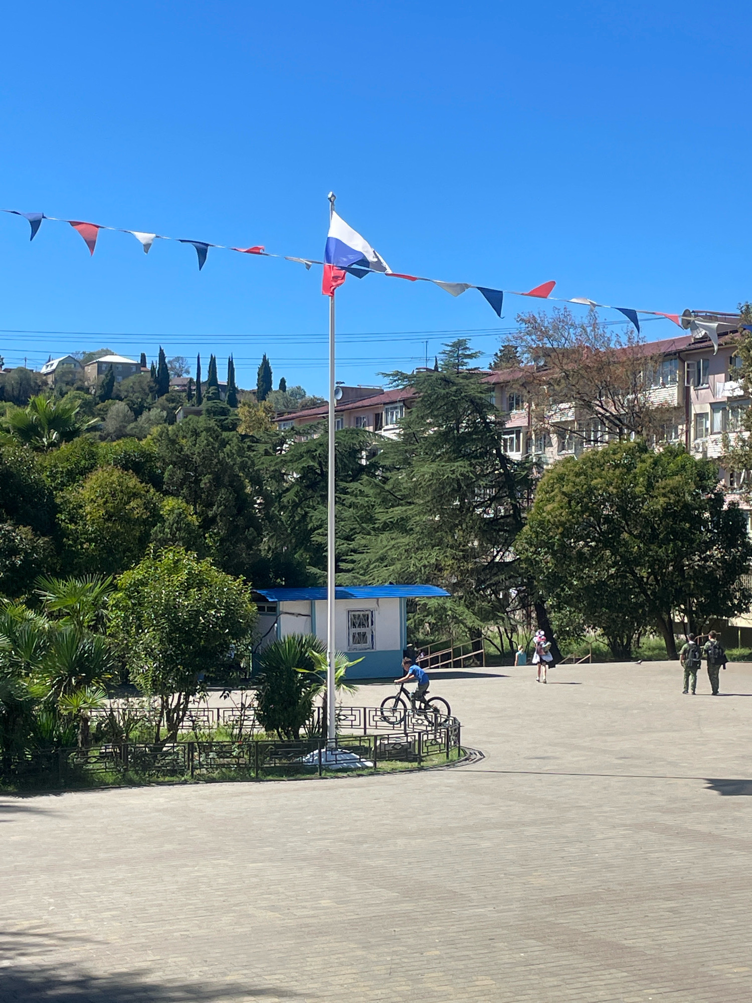In Sochi, Russia, on a sunny day with a clear blue sky, a courtyard area is depicted. A Russian flag flies on a flagpole in the center of the scene, with a string of red, white, and blue triangular flags draped across the sky. In the foreground, a boy on a bicycle rides near a low fence and some greenery. Further back, two people walk away from the viewer, and another person is seen in the distance. Several buildings and lush green trees surround the courtyard, with a hill rising in the background.