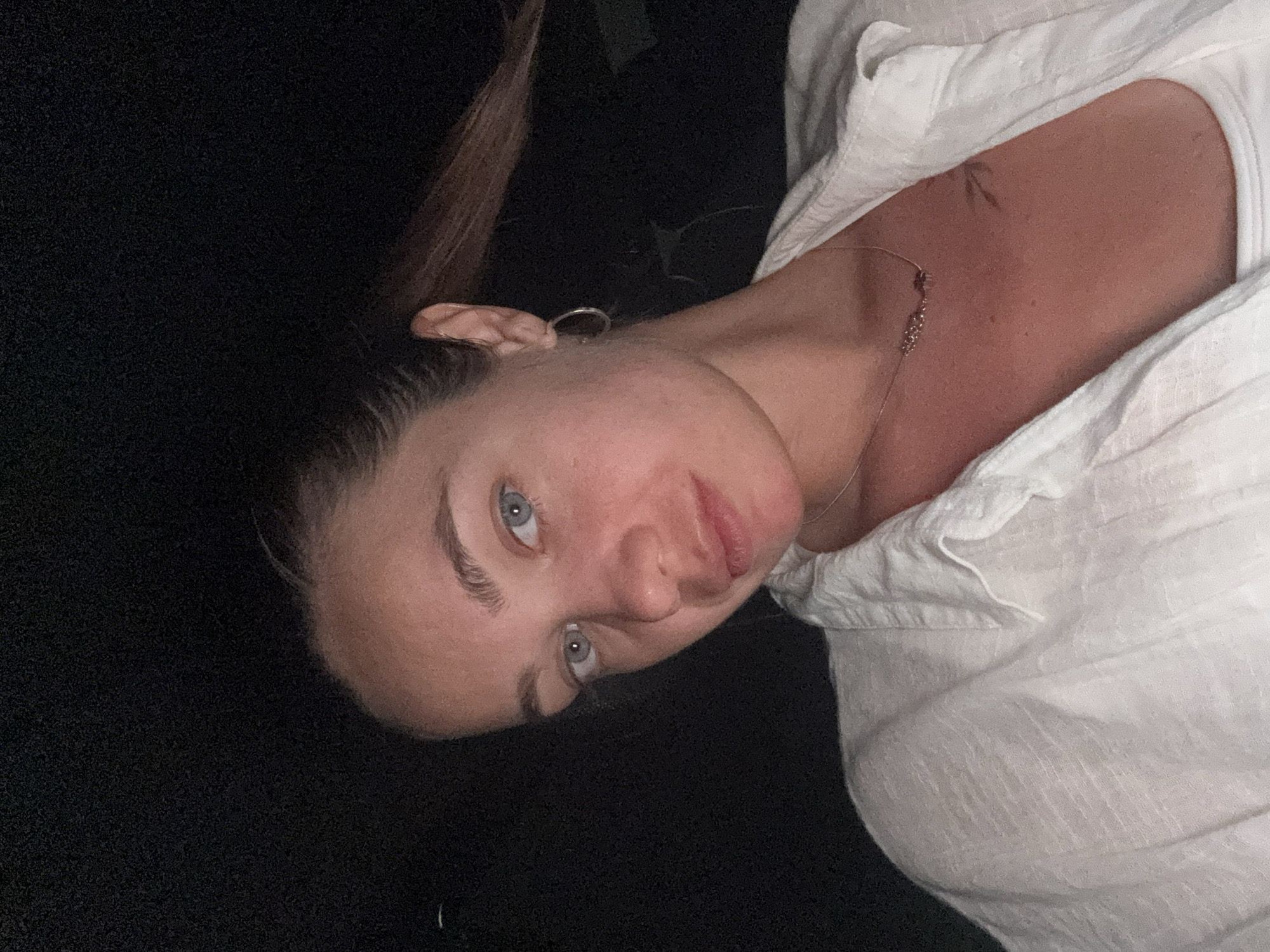A young woman with light blue eyes and light brown hair pulled back in a ponytail is pictured in a close-up selfie. She is wearing a white linen shirt. Her left ear is adorned with a silver hoop earring. A delicate silver necklace rests on her collarbone. The background is dark, suggesting an evening or indoor setting, with no discernible city landmarks of Radovići, Montenegro. The lighting focuses on her face and upper torso, highlighting her skin tone.