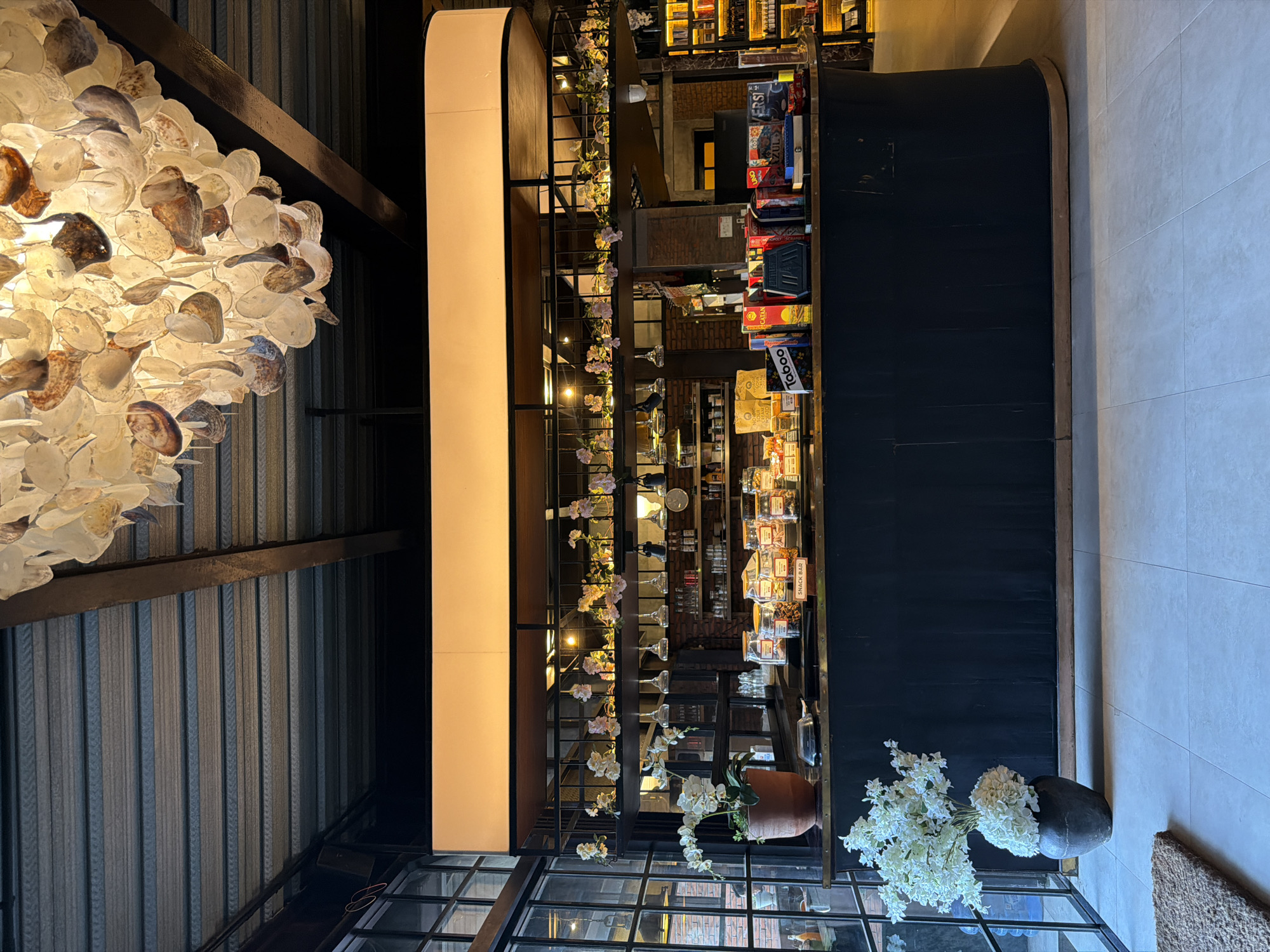 This is a low-angle, eye-level shot of a dimly lit, modern cafe counter area. The counter is dark wood with a brass trim. On top of the counter, there are several glass jars filled with snacks, some boxes of games, and a sign that reads "SNACK BAR". To the left of the counter, there is a large vase of white flowers. Behind the counter, there are shelves stocked with various items. The ceiling is made of exposed beams and metal sheeting, and a large, ornate chandelier hangs from it. The cafe appears to be located in North Kuta, Indonesia, based on the context provided. There are no people visible in the image, and no actions or interactions are taking place.