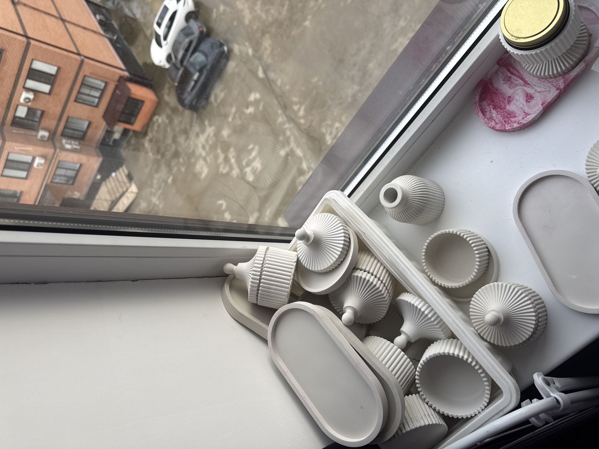The image shows a window sill with a collection of decorative items. The items are primarily white and appear to be made of a material like plaster or ceramic. There are several small vases, decorative trays in various shapes (oval, round), and objects with a ribbed or fluted design. One item has a gold-colored lid.
Outside the window, there is a view of a building with orange-colored bricks, and what appears to be a reflection in a puddle or wet surface. Two cars are visible, one white and one grey. The location is unspecified, but the scene suggests an urban environment.