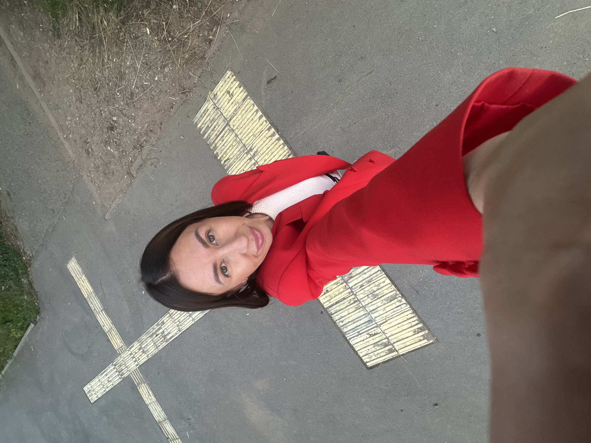 An overhead, first-person perspective selfie shows a woman in a red blazer standing on a paved surface with tactile paving indicators. The woman has dark hair and is looking at the camera with a slight smile. She is wearing a white top under the blazer. The tactile paving is yellow and appears to be a pedestrian crossing or warning surface. The environment is outdoors, with some dry grass and dirt visible to the right. The scene is likely a street or sidewalk in the city of Votkinsk, Russia, given the presence of tactile paving which is common in Russian cities for accessibility.