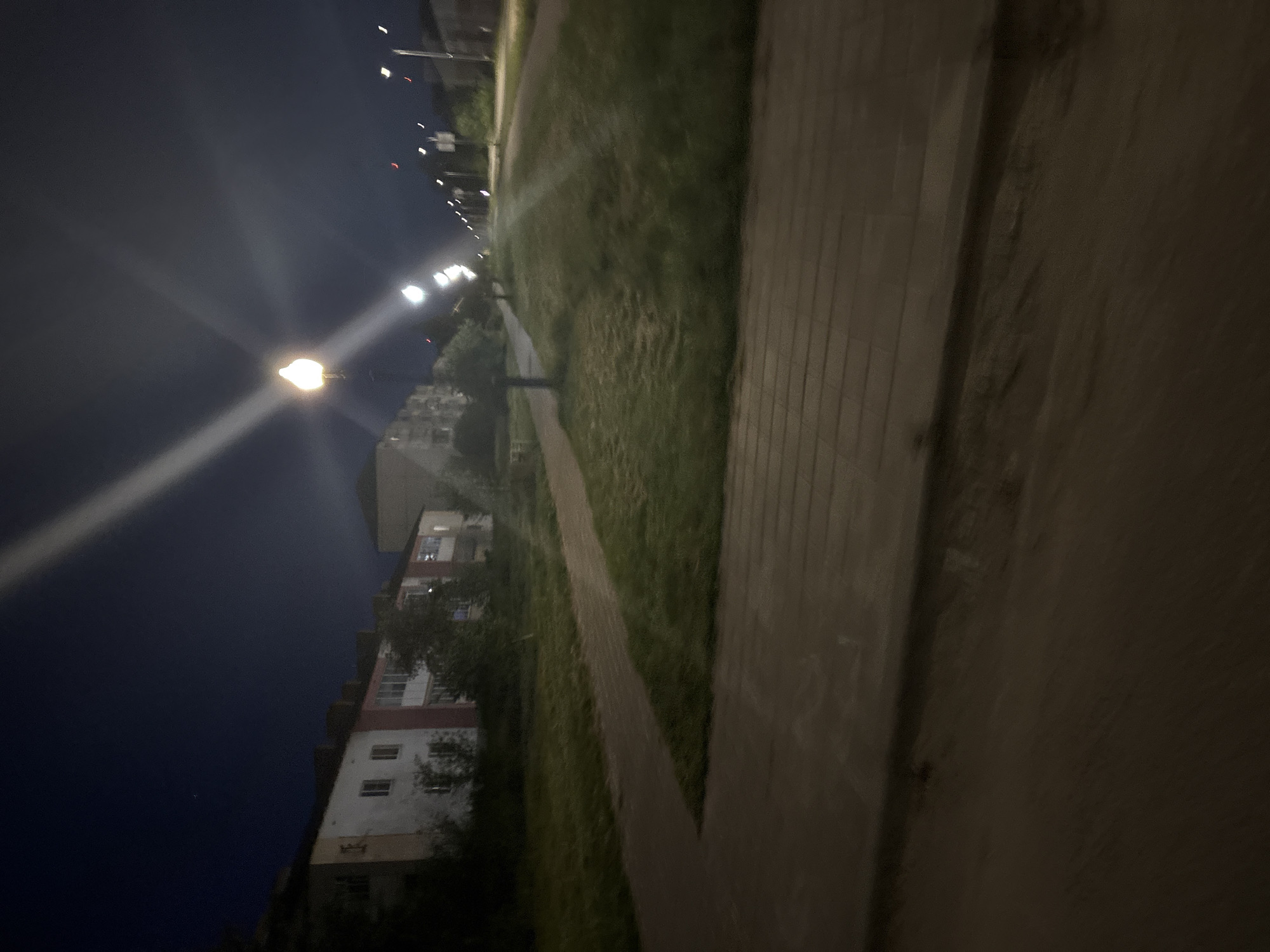 A nighttime outdoor scene captured in the city of Unknown, Unknown, presents a quiet residential or suburban environment. The sky is a dark blue-black with a single faint white point visible in the upper left quadrant. On the left side of the image, a row of multi-story buildings, likely residential, exhibits white and reddish-pink facade sections with visible windows, some dimly lit from within. These structures are partially obscured at their base by dark green foliage from trees and bushes.
In the midground, a paved pathway composed of rectangular dark grey and brown pavers runs parallel to the buildings, curving slightly to the right as it recedes. Adjacent to this pathway on the right is a strip of overgrown green grass. Further to the right, a wider expanse of unkempt grass leads toward a distant road. A prominent streetlight in the mid-right foreground emits a bright yellow-white light, creating a significant lens flare that extends diagonally across the upper portion of the image. A series of additional streetlights, visible in the distance along the road, also produce distinct bright spots and lens flares. The immediate foreground is dominated by a dark, textured surface, appearing to be pavement or a roadway. No individuals, animals, or vehicles are present in the scene.