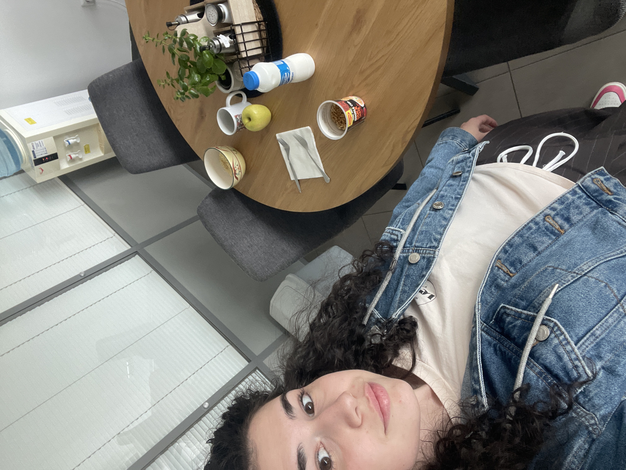 The photograph appears to be a selfie taken indoors, possibly in an office or co-working space. The focal point is a woman with curly dark hair wearing a light-colored t-shirt and a denim jacket. She is oriented towards the camera at an angle.
In the background is a round wooden table. Objects on the table include a plant, a spice rack, a bottle of milk, a mug, an apple, a bowl, a cup of instant noodles, and utensils on a napkin. There are grey chairs around the table.
To the left, there is a water cooler. The floor appears to be tiled. In the background, there is a glimpse of a sofa and a shoe.
Without additional information, it is not possible to definitively confirm the location as Sofia, Bulgaria.