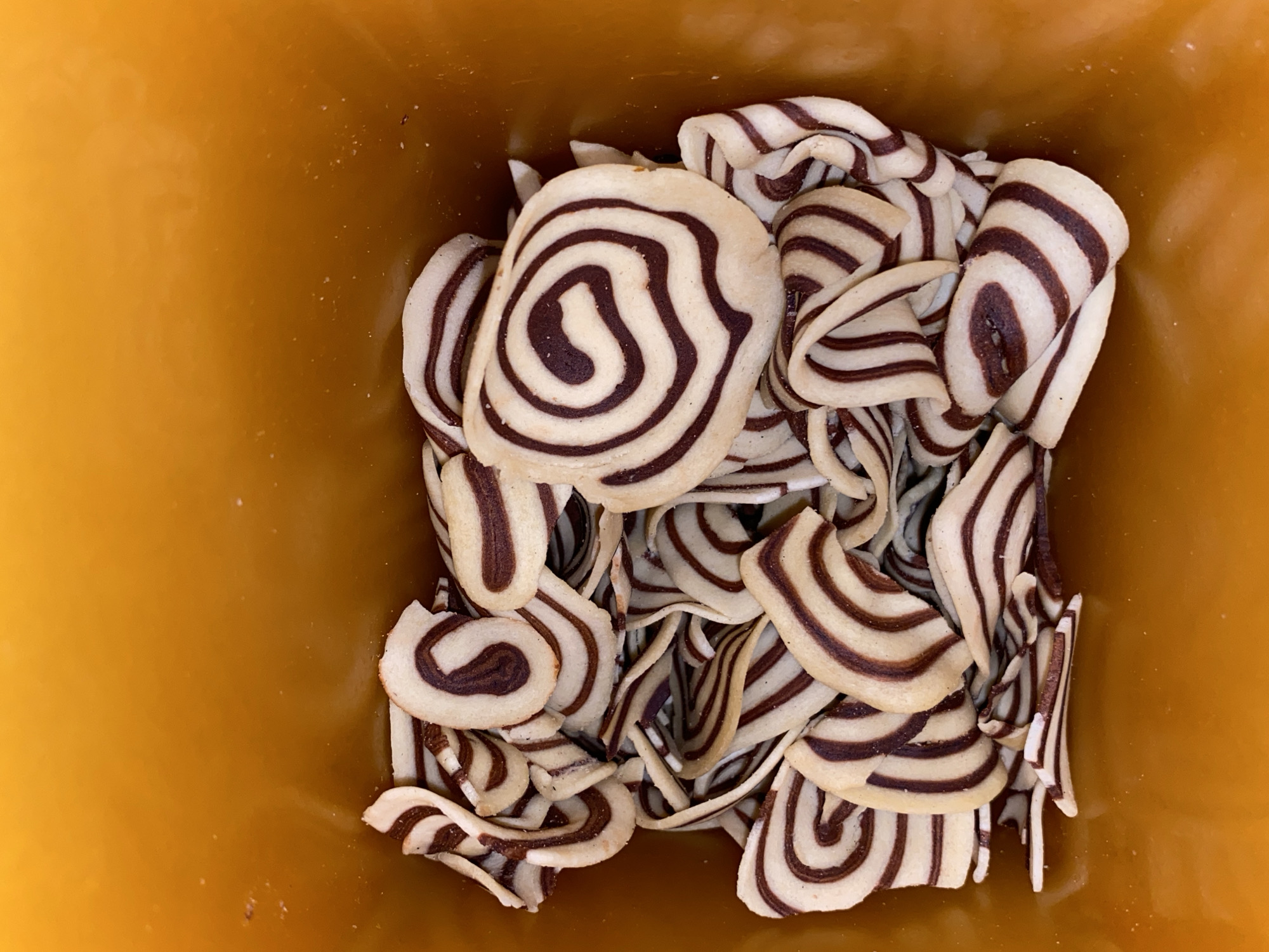 The image shows a container filled with spiral-patterned cookies. The cookies are generally circular, with alternating light and dark brown swirls. The container is a solid, light brown-orange color. The image appears to have been taken from directly overhead. There are no people in the image. The location is not indicated within the image, so it is impossible to verify if it is in Lhokseumawe, Indonesia.