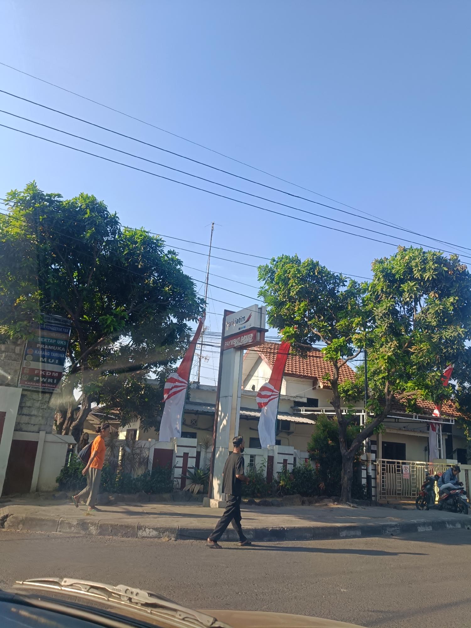 A street scene in Keboansikep, Indonesia shows a paved road in the foreground with a sidewalk adjacent to it. On the sidewalk, two individuals are walking. The person on the left is wearing an orange shirt and grey pants, carrying a blue bag over their shoulder. The person on the right is wearing a black shirt and black pants. In the background, there is a building with a red-tiled roof and white walls, adorned with two Indonesian flags. A sign with Indonesian text is visible on a pole in front of the building, next to a large green tree. Further to the right, two motorcycles are parked, with one person sitting on one of them, looking at their phone. The sky is clear blue with several overhead power lines crossing the frame.