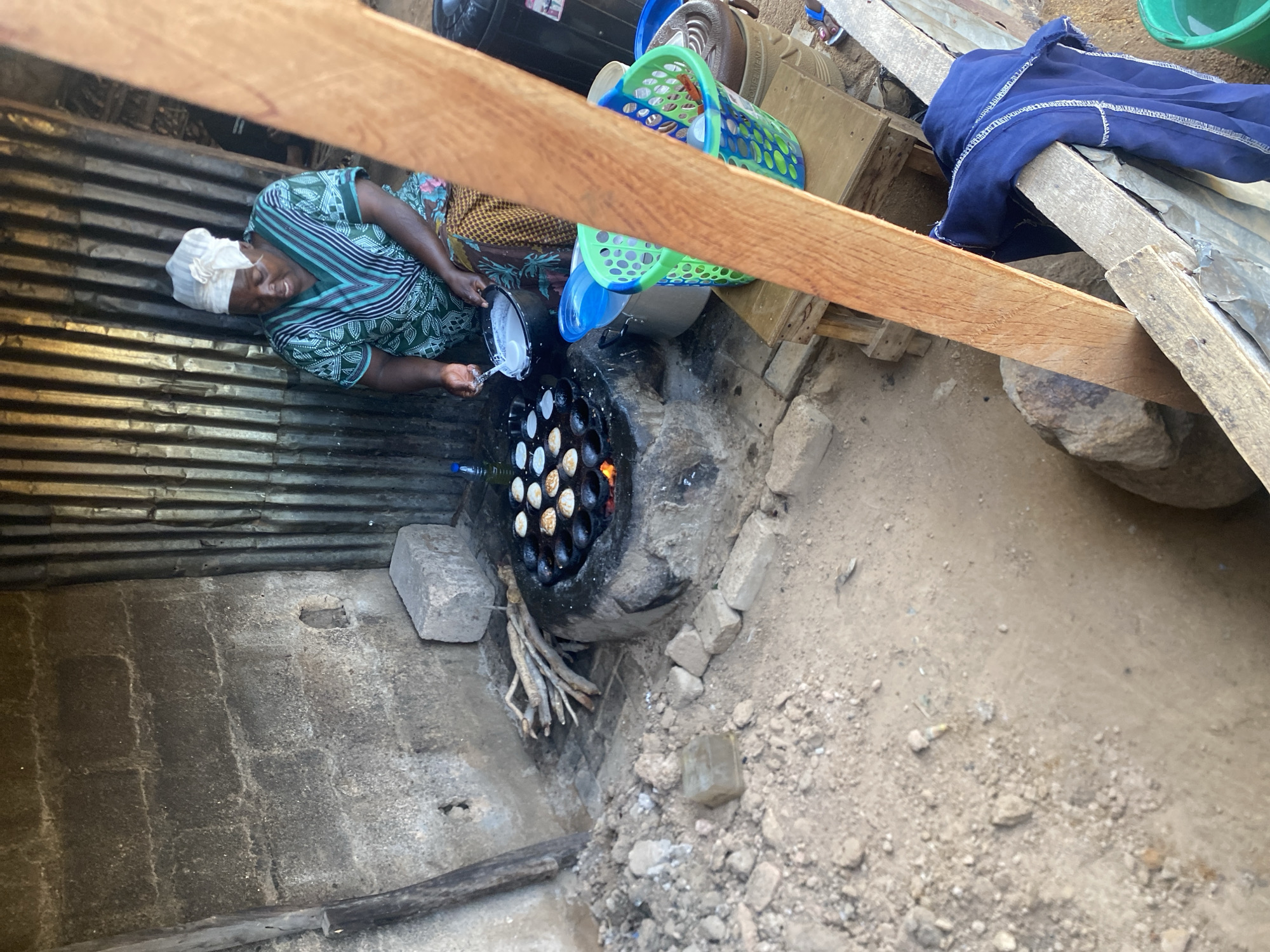 In the city of Jos, Nigeria, a woman is seen cooking what appear to be small pancakes over an open fire. She is wearing a headwrap and a patterned green and white shirt, and she is pouring batter from a white bowl into a black, multi-holed griddle. The griddle is set into a rough, stone-lined pit that is heated by burning wood. Scattered around the cooking area are piles of dirt, rocks, and various cooking utensils, including a green and blue plastic basket containing more bowls. A corrugated metal wall forms the backdrop of the scene. The overall environment suggests an outdoor, informal cooking setup.