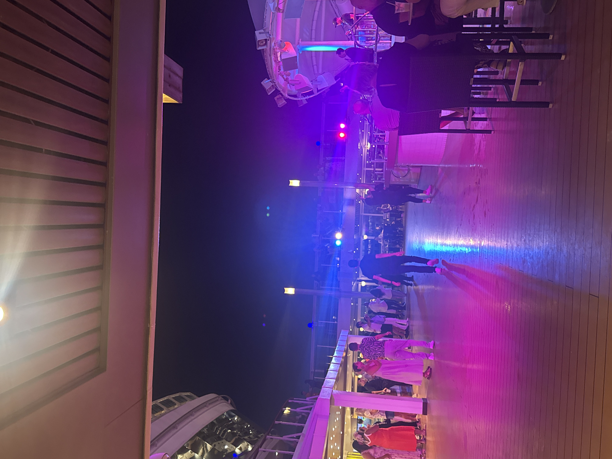 The image depicts an outdoor nighttime scene with a focus on a lively gathering. Multiple individuals are visible, some standing and conversing, while others appear to be walking or dancing. The environment is illuminated by vibrant stage lighting in shades of pink, blue, and purple, casting colorful reflections on the wooden floor. There is a stage or performance area in the background with speakers and lighting equipment. The architecture suggests a modern open-air venue, possibly a cruise ship deck or a resort area. The overall atmosphere appears festive and social. The location of Ibiza, Spain is not evident from the visual information provided.