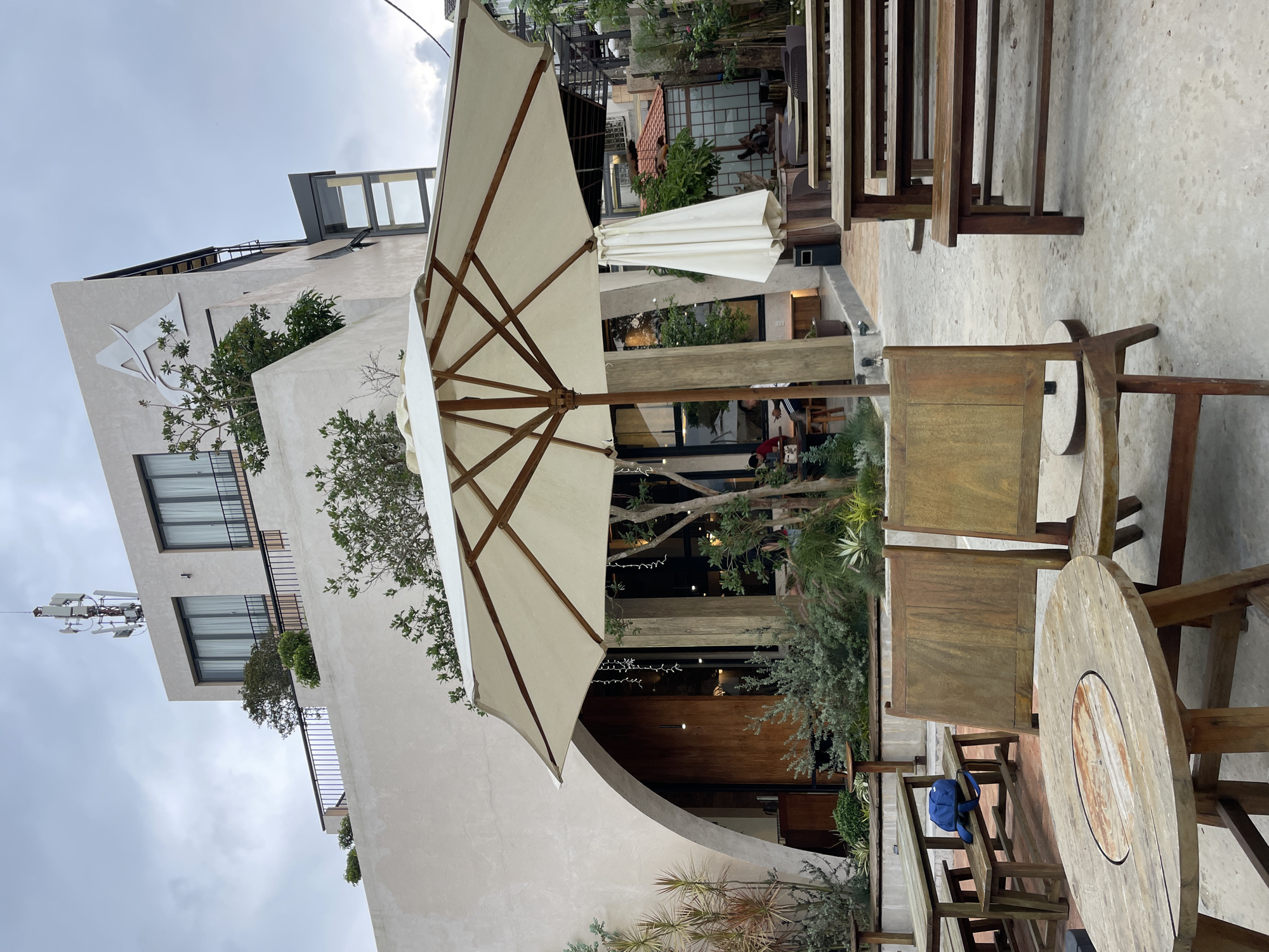 The image captures an outdoor seating area of a cafe in Da Lat, Vietnam, under an overcast sky. In the foreground, a large white umbrella is open, supported by a wooden pole. Two wooden chairs sit at a round wooden table with a rustic top. Behind these, more wooden benches and tables are arranged. The cafe building, with its modern, off-white facade and an abstract logo at the top, dominates the background. Large windows reveal glimpses of the interior, where a few patrons can be seen. Lush greenery, including potted plants and trees, is integrated into the cafe's design, softening the architectural lines. The overall atmosphere suggests a relaxed, contemporary cafe setting in Da Lat's cooler climate.