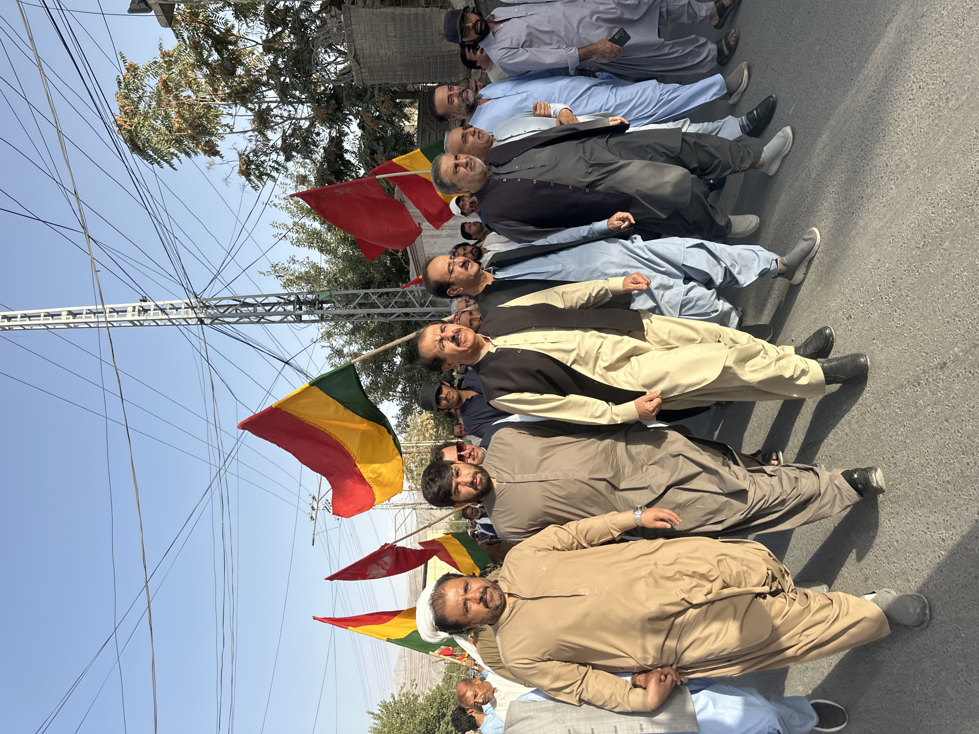 A group of men are walking in a procession on a paved road in Quetta City Tehsil, Pakistan. They are holding several flags, including a large tricolor flag of red, yellow, and green, and a red flag. The men are dressed in traditional Pakistani clothing, such as shalwar kameez and vests. Some are wearing caps, and one man is wearing sunglasses. The background shows a few trees and a utility pole with many wires. The sky is clear and blue. The scene appears to be a political rally or demonstration.