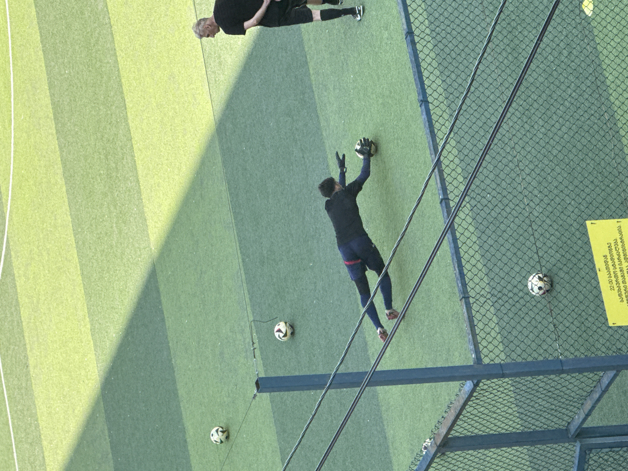 The image shows a soccer field with two men. One man, wearing a black shirt and black shorts, is standing and watching. The other man, wearing a dark long-sleeved shirt and dark pants, is in the process of diving to save a soccer ball. The field is made of green artificial turf with darker green stripes. There are several soccer balls visible on the field. In the foreground, there is a chain-link fence and some wires. A yellow sign with Georgian text is visible on the fence. The scene is outdoors, and sunlight is casting shadows on the field. This appears to be a training or practice session.