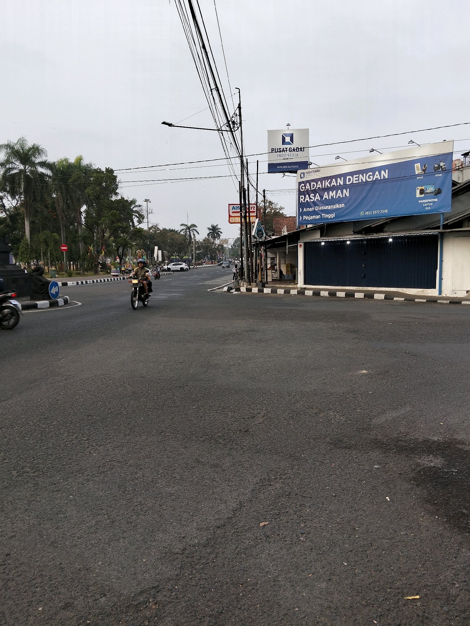 The image depicts a street scene in Purworejo, Indonesia, likely near a commercial area. In the foreground, a gray asphalt road shows signs of wear and tear, with faint markings on the surface. A white line demarcates the lane, and a black and white checkered pattern runs along the edge of the road, indicating a pedestrian crossing or median.
Several vehicles are present. A motorcycle with a rider wearing a helmet is prominently positioned in the intersection, moving forward. Further down the road, more vehicles, including cars and at least one other motorcycle, are visible. The presence of these vehicles suggests a moderately busy urban environment.
On the right side of the road, a building with a dark blue corrugated metal facade is visible, likely a shop or commercial establishment. Above the building, a large blue banner displays text in white and yellow, advertising a service that involves pawning items like gold, laptops, and cameras. The banner includes contact information, a phone number: "0811 1972 2548". To the left of this building, another smaller billboard with a white background and blue text reads "PUSAT GADAI INDONESIA" and "MAJEN SUTOYO".
The environment is somewhat overcast, with a pale sky. Lush green palm trees and other foliage line the left side of the street, suggesting the presence of a park or green space. Streetlights are mounted on poles, and an intricate network of overhead power lines crisscrosses the sky, a common sight in urban Indonesian landscapes. A red circular traffic sign with a white horizontal bar is also visible on the left side of the road. The overall impression is of a typical day in an Indonesian city, with everyday activities and commercial advertising dominating the scene.