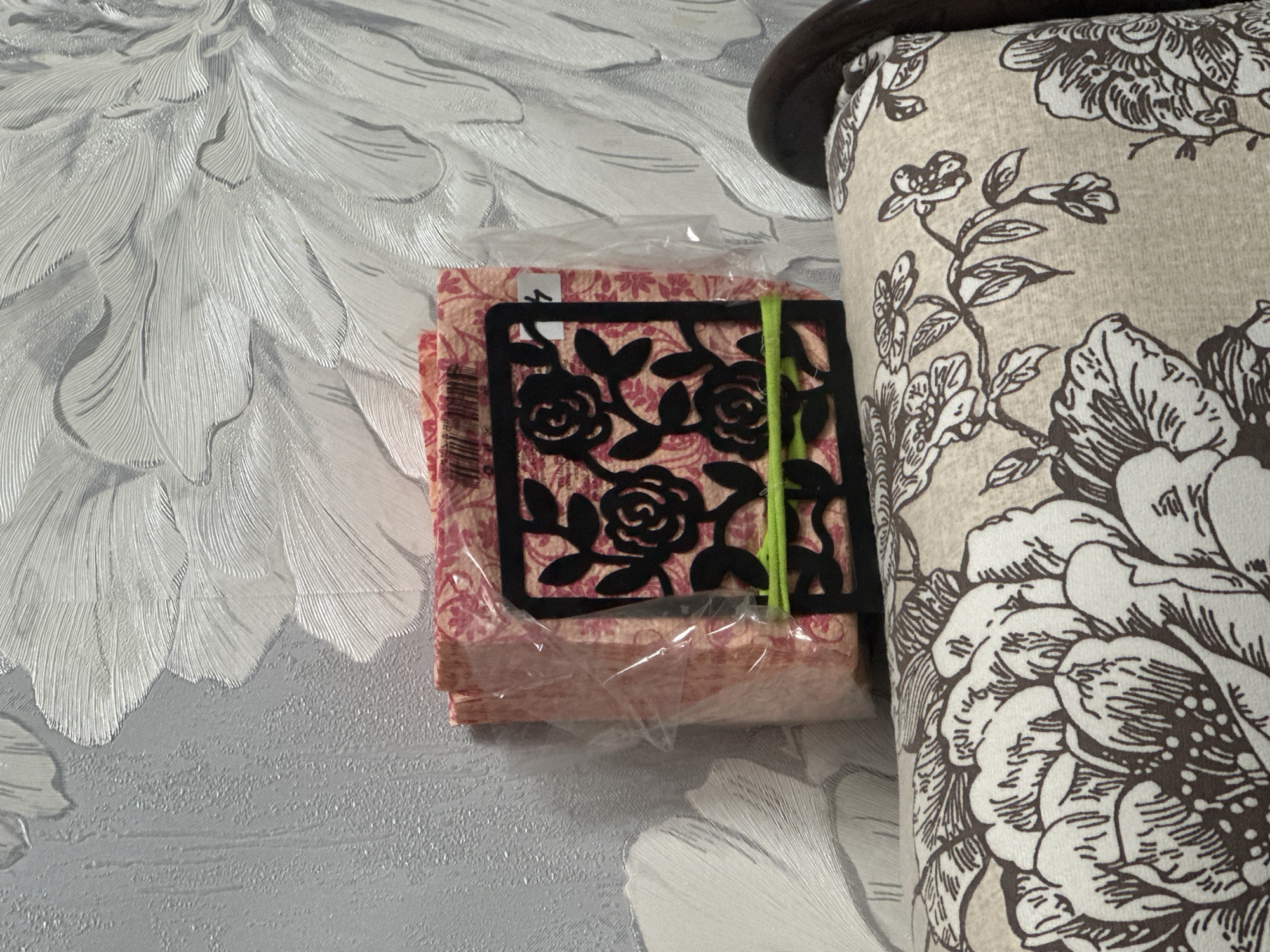 A close-up shot captures a stack of folded fabric napkins in a plastic wrapper, held together by a bright green elastic band. A black metal bookmark with a rose design is placed over the napkins. The background features a textured, pale grey wallpaper with a large, embossed floral pattern. To the right of the napkins, a portion of a sofa or armchair is visible, upholstered in a light beige fabric with a darker brown floral print. The lighting suggests an indoor setting, possibly in daylight, casting soft shadows. The overall impression is domestic and decorative. No people are visible, and the focus is on the arrangement of objects and the surrounding textures and patterns. The image is taken from a slightly elevated perspective, looking down at the items.