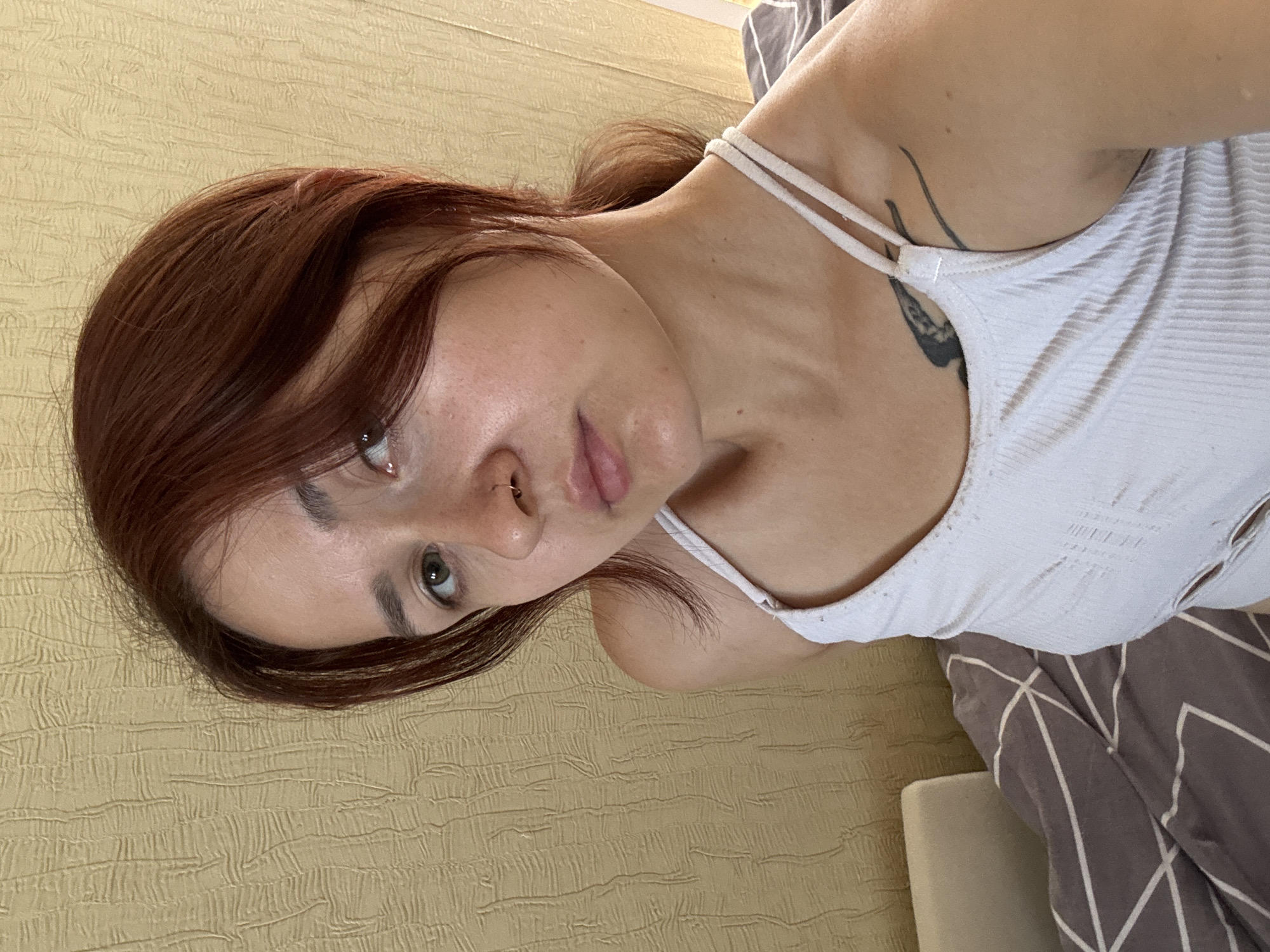 A young woman with auburn hair, fair skin, and green eyes is shown in a close-up, high-angle selfie. She is wearing a white ribbed tank top with thin straps and a nose ring. A tattoo is visible on her left shoulder. She is positioned in front of a textured, yellowish wall. To her left, a portion of a bed with a gray and white patterned comforter is visible. The lighting suggests an indoor setting, likely during the daytime. No specific location within Naberezhnye Chelny, Russia, can be determined from the visual information.