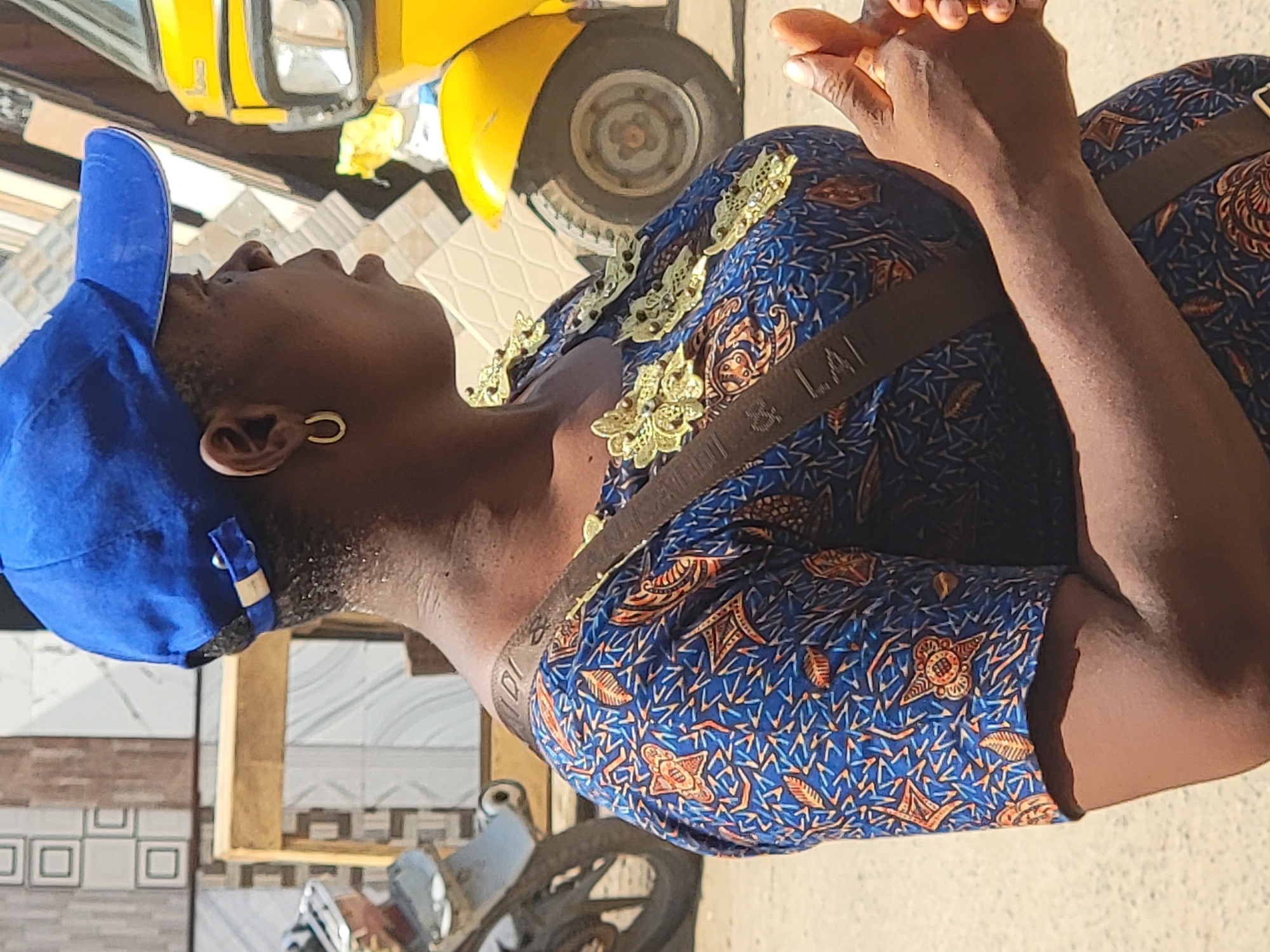 This vertical, brightly lit outdoor shot captures a person in profile, looking upwards with hands clasped together, suggesting a moment of prayer or contemplation. The individual wears a blue patterned garment adorned with gold-colored, leaf-like appliqués on the shoulder area, and a blue baseball cap worn backward. A single gold hoop earring is visible in their ear. A brown strap, possibly from a shoulder bag, crosses their body, with the partial text "LAI" visible on it.
In the background, a bright yellow vehicle, likely a tricycle taxi (keke napep) common in Nigeria, is partially visible at the top, along with one of its wheels. The ground below features various patterned tiles, and a wooden pallet or structure is also present. Part of a motorcycle or bicycle can be seen towards the bottom. The strong overhead sunlight indicates it is daytime, possibly midday, casting clear shadows and highlighting textures. The scene is set in an outdoor, likely urban or semi-urban environment in Maiburiji, Nigeria.