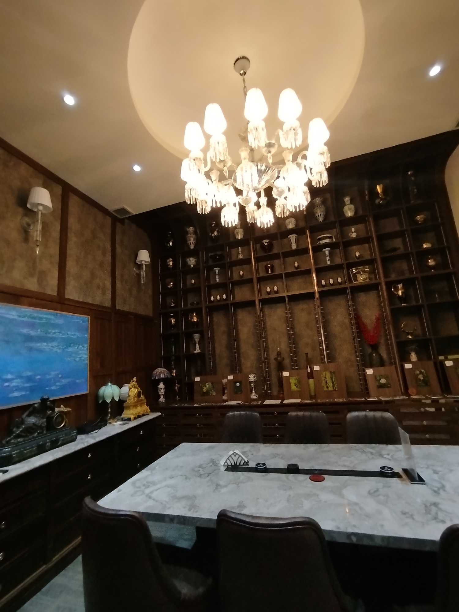 The image depicts an indoor scene in what appears to be a room with ornate decor. In the foreground, a marble-topped table with a dark base is visible, with several dark leather chairs around it. The table has a central black organizer with circular elements and a red accent. In the background, a large wooden shelving unit spans across the wall, filled with numerous decorative items, including vases and other collectibles. To the left of the shelving unit, a large painting with blue and white hues hangs on a wood-paneled wall. Above the painting, a wall-mounted lamp with a white shade is visible. The ceiling features a large, recessed circular cove with a prominent crystal chandelier hanging from its center, casting light on the room. The overall ambiance suggests a well-decorated space, possibly a dining room or a study. There are no people visible in the image. The location is identified as Phường Xuân Hương - Đà Lạt, Vietnam.