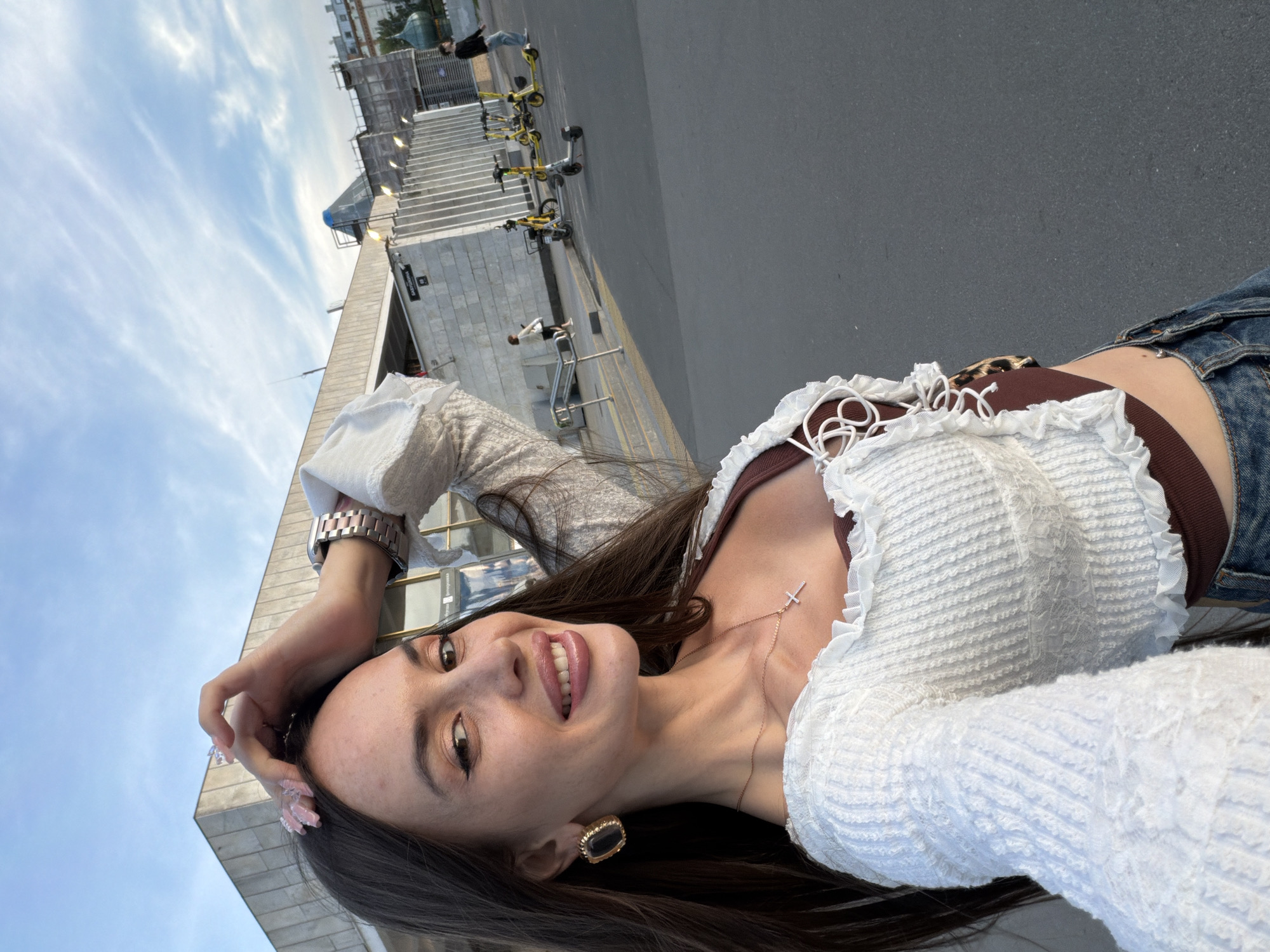 A young woman with long dark hair, wearing a white ruffled top over a brown bralette and blue jeans, takes a selfie outdoors. She is wearing large gold earrings and a silver necklace with a cross pendant. Her left hand is raised and resting on her head, and she is smiling, with her tongue slightly protruding. In the background, a modern building with a concrete facade is visible, along with several yellow electric scooters parked on the pavement. The sky is cloudy with streaks of blue. The overall scene suggests an urban environment on a bright day.