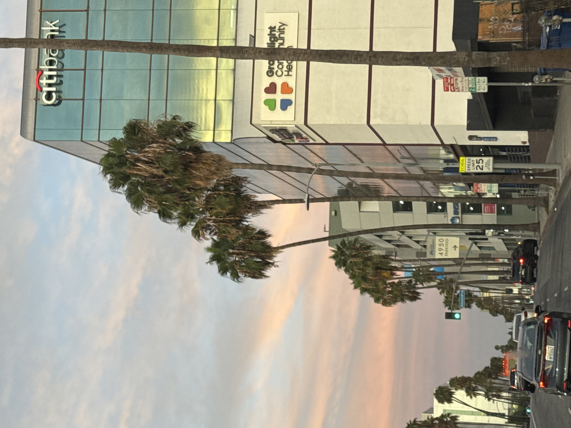 This image depicts a street scene in Los Angeles, California, USA, at dusk. A multi-story building with a glass facade, featuring the "Citibank" logo, dominates the right side of the frame. Adjacent to it is another building with a white exterior, displaying the "Graylight Community Health" logo. Palm trees line the street, casting shadows and adding to the urban tropical ambiance. Several cars are visible on the road, some in motion and others parked. Traffic signals and street signs, including one indicating "4950 PARKING" with an arrow and another for "SCHOOL SPEED LIMIT 25," are also present. The sky is painted with soft hues of pink and orange, indicative of a sunset. The overall scene conveys a typical urban environment with a blend of commercial buildings, traffic, and natural elements.