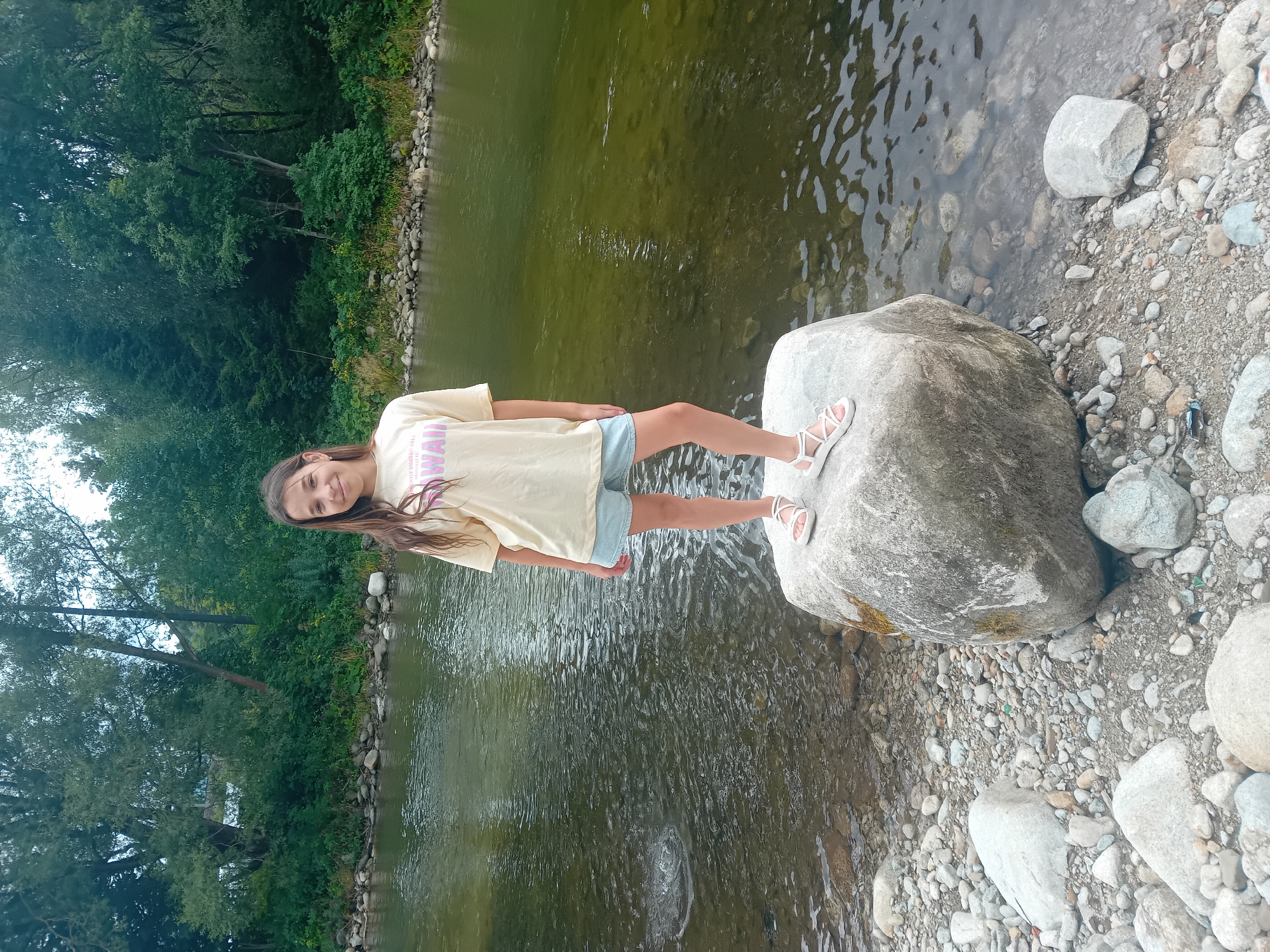 A young girl with long brown hair, wearing a light yellow t-shirt and denim shorts, stands on a large rock by a river. She is smiling at the camera. The river water is clear with ripples and reflections of the trees on the opposite bank. The riverbank is covered with stones of various sizes, and lush green trees form a dense backdrop. The scene appears to be a natural outdoor setting, possibly in the city of Habovka, Slovakia, given the context provided.
