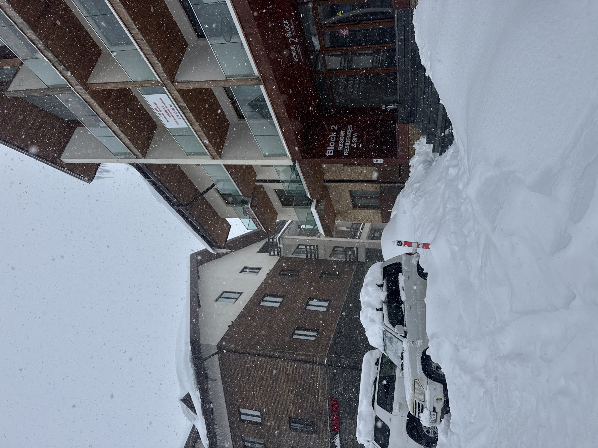 The image shows a snowy scene in what appears to be a resort area. Several buildings are visible, including a building identified as "Block 2 Resort Residences & Spa." Snow is heavily present, covering vehicles parked nearby. The scene is likely outdoors, with a wintry, snowy atmosphere. There is some text in Cyrillic on a building.