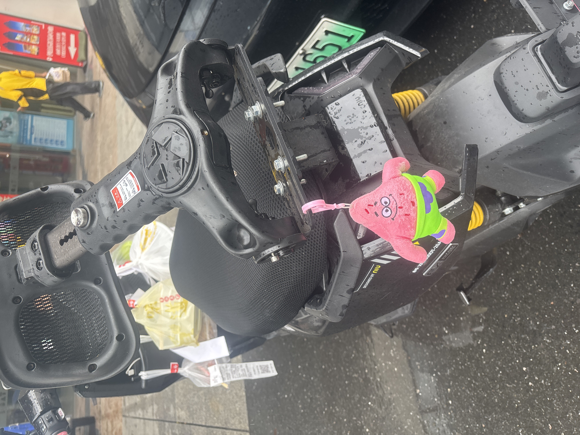 In Baiyun District, China, a close-up shot captures a black electric scooter parked on a wet street, suggesting recent rainfall. A stuffed toy of Patrick Star from SpongeBob SquarePants, wearing green shorts, is hanging from the scooter's rear. The scooter is adorned with various accessories, including a basket at the front, and a license plate with the number "1651" is visible on the back. A person in a yellow jacket and dark pants is walking in the background, carrying shopping bags. The scene is set in an urban environment with shopfronts visible.