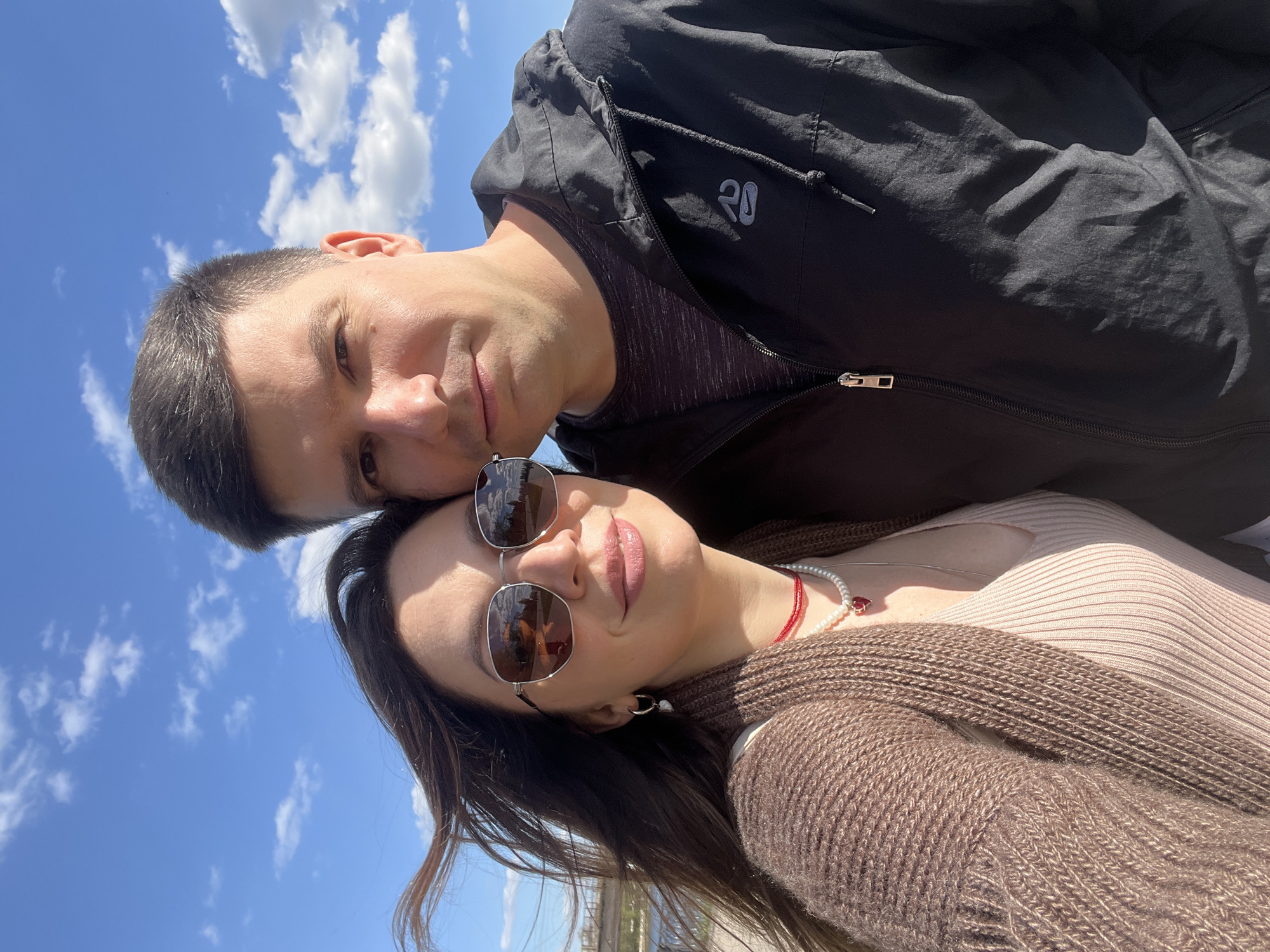 A selfie-style photograph shows a man and a woman close together against a bright blue sky with scattered white clouds. The man is on top, wearing a black windbreaker jacket with a logo on the left chest. He has dark hair and a neutral expression. The woman is beneath him, wearing sunglasses and a brown sweater. She also has dark hair and a light-colored top underneath the sweater. She is wearing a necklace with red and white beads. The background suggests they may be outdoors in an urban area due to the presence of a structure near the bottom left of the image. The city is unknown.