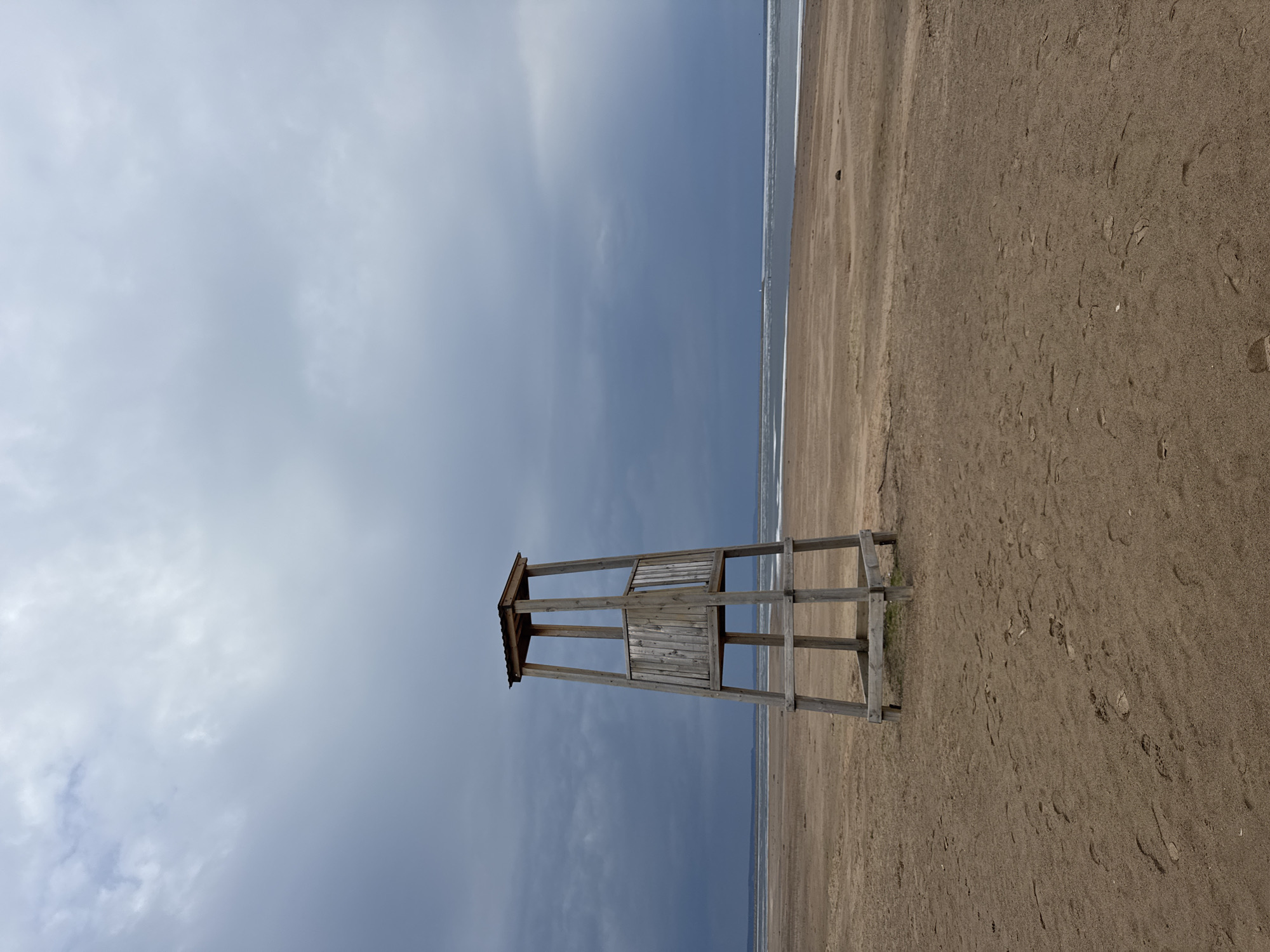 The image depicts a desolate beach scene in Chaikovsky, Russia. A weathered, wooden lifeguard tower stands alone on the sand, its slats partially blocking the view of the ocean. The sky is overcast with a mixture of light and dark clouds. The sand is a light brown color and there are a few footprints visible. The ocean is a narrow strip of water in the background. There are no people visible in the image.