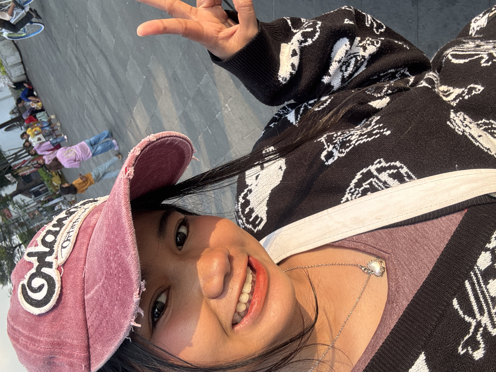 A young woman wearing a pink baseball cap and a black cardigan with white patterns is taking a selfie outdoors. She is smiling and making a peace sign with her right hand. In the background, several children are playing and sitting on the ground in what appears to be a public square. The ground is paved with grey tiles. The lighting suggests it is daytime, possibly late afternoon, with warm sunlight casting shadows. The scene is likely located in a city square or park within West Jakarta, Indonesia, as indicated by the presence of street vendors and local attire often seen in such locations in the region.