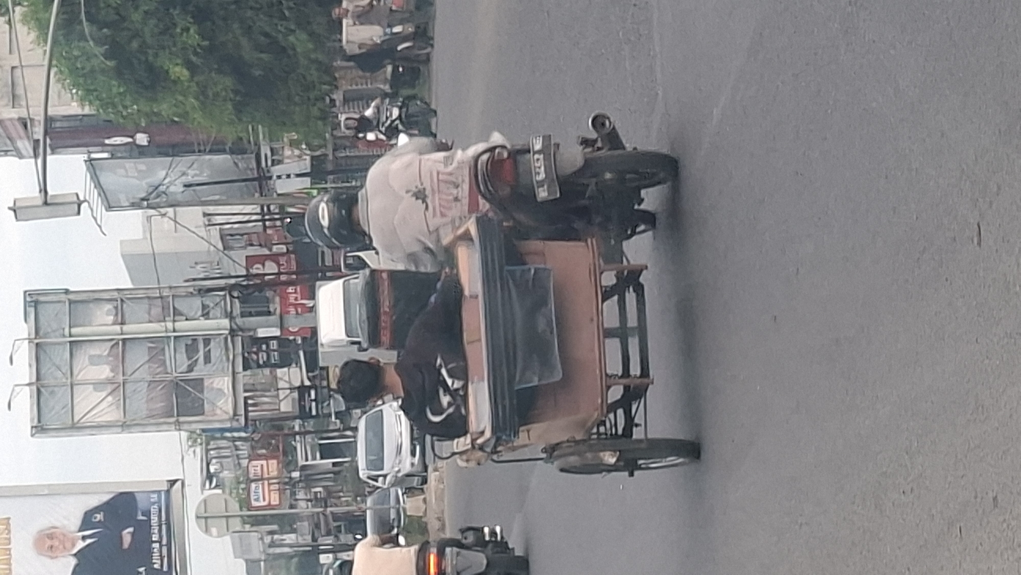 In Lhokseumawe, Indonesia, a sidecar motorcycle with two occupants is traveling on a paved road. The driver, wearing a light-colored jacket and a helmet, is operating the motorcycle. Behind him, a passenger is seated in the sidecar, wearing a dark hooded sweatshirt. The sidecar is a brown, box-like structure with a black tarp covering part of it. The motorcycle has a license plate that reads "BL 6452 WG". In the background, there are several buildings, billboards, and other vehicles, suggesting an urban environment. One billboard features a portrait of a man in a suit. The sky is overcast.