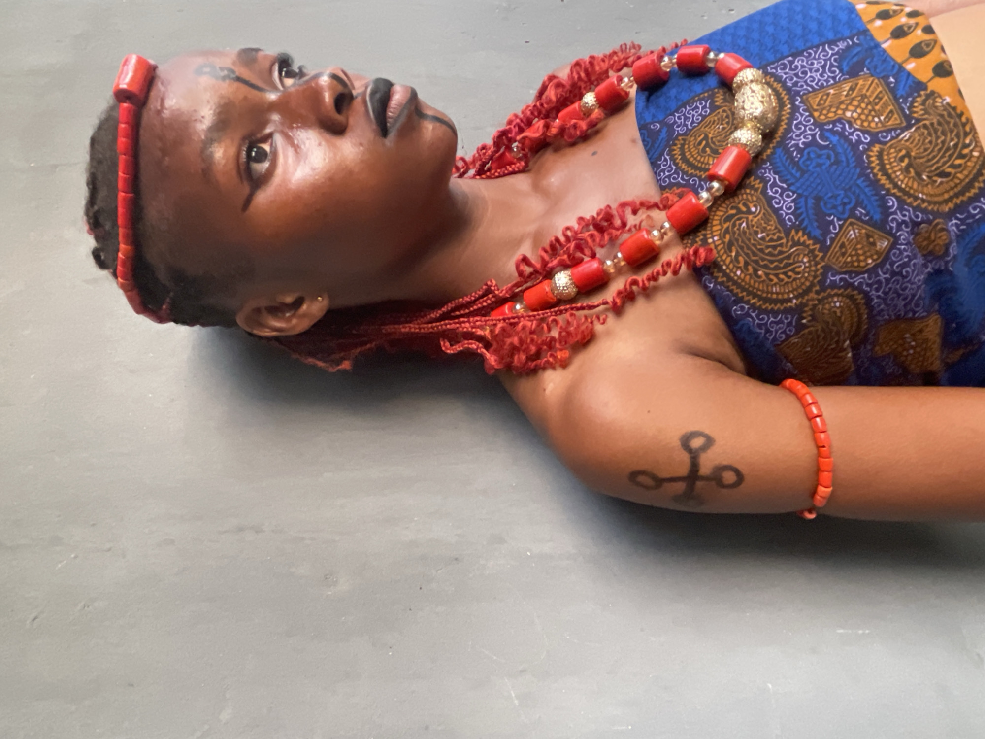 The image portrays a person, likely a young woman, lying on a light grey, flat surface, possibly concrete. She is positioned looking upwards and slightly to her right, with a calm and contemplative expression.
She is richly adorned in what appears to be traditional attire and accessories. A red beaded headband, possibly made of coral, frames her face. Around her neck and upper chest, she wears an elaborate multi-strand necklace of red coral beads, interspersed with decorative gold-colored metallic beads and clear spacers. Red, textured fabric or tassels are also visible around her neck area, possibly part of the necklace or her garment.
Her body is partially covered by a vibrant garment featuring a blue, gold, and brown pattern, characteristic of African textiles such as Ankara. On her right forearm, she wears a matching red beaded bracelet. A distinct design, resembling a cross with circular elements at the ends of its arms, is visible as a tattoo or body paint on her upper right arm. Her face features black linear markings on her forehead and extending downwards from her lower lip. She is wearing black lipstick and a small gold earring is visible in her left ear. Her hair is styled in short, dark braids or cornrows.
The minimalist background emphasizes the subject and her detailed adornments. The lighting is soft and even, suggesting an indoor or shaded setting. The overall aesthetic, including the coral beads, patterned fabric, and facial markings, points to a strong cultural or ceremonial context. Given the location context of Badagry, Nigeria, these elements align with the rich cultural heritage, particularly of the Yoruba and Edo people, where such adornments signify status, tradition, and identity. The pose suggests a staged photograph, possibly for artistic expression or documentation of cultural practices.