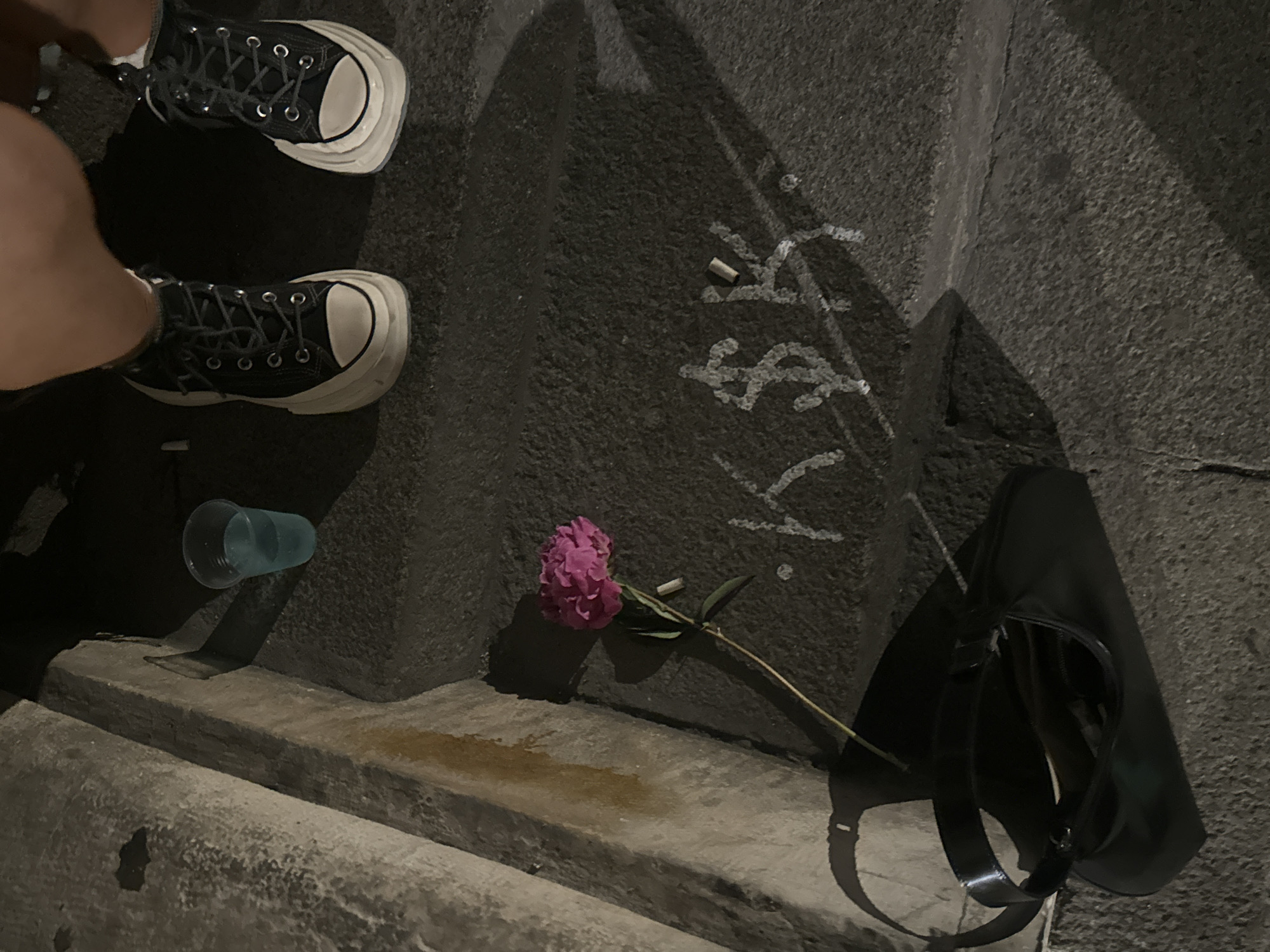 The image depicts a low-light scene on a series of textured concrete or stone steps, reportedly located in Sofia, Bulgaria. In the upper right portion, the lower legs and feet of an individual are visible, wearing black high-top platform Converse sneakers with white soles and toe caps. To the left of the footwear, a clear plastic cup holds a small volume of light blue liquid on a step. Approximately centered on a lower step, white chalk-like markings spell "K $ K," preceded by a dot and followed by a partial character. A single cigarette butt is positioned vertically between the dollar sign and the second 'K'. A vibrant pink peony flower, featuring a green stem and leaves, rests diagonally across the steps towards the lower-left. On the lowest visible step in the bottom-left quadrant, a black shoulder bag or purse with a glossy strap is lying on its side. The steps show signs of wear and minor discoloration, and strong shadows are cast across the surfaces due to limited illumination.