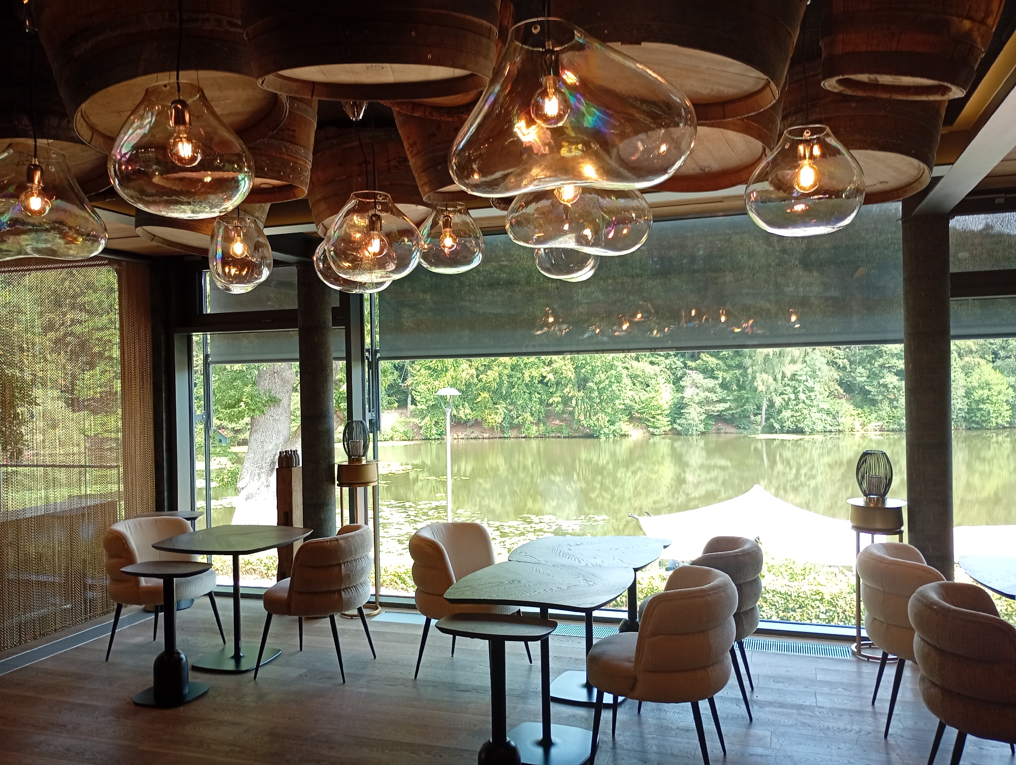 The image depicts the interior of a restaurant or lounge area with a view of a tranquil lake and lush greenery. The room features a modern, sophisticated design with exposed wooden barrel halves on the ceiling, from which several large, bulbous glass pendant lights with visible Edison-style bulbs hang. The lighting is warm, casting a golden glow throughout the space.
The seating area consists of several tables and chairs. Some tables are dark, oval shapes with slender legs, while others are smaller, circular side tables. The chairs are upholstered in a textured, neutral fabric, likely beige or cream, with a ribbed design on the backrests and seats. They have dark, angled legs.
Large windows dominate the back wall, offering an expansive view of a calm lake surrounded by dense green trees. Lily pads are visible on the water's surface. In the middle ground, across the lake, a white tent-like structure is partially visible.
The flooring is a dark wood laminate. Decorative elements include tall, cylindrical lamps with ornate, cage-like shades on pedestal bases placed near the windows, and a dark sculptural object on a wooden stand. A gold-colored chain-link curtain is also present on the left side of the image. The overall impression is one of a chic, relaxing establishment with a connection to nature. This scene is likely located at "Waldsee Restaurant & Bar" in Bad Sachsa, Germany, given the distinctive view of the Waldsee.