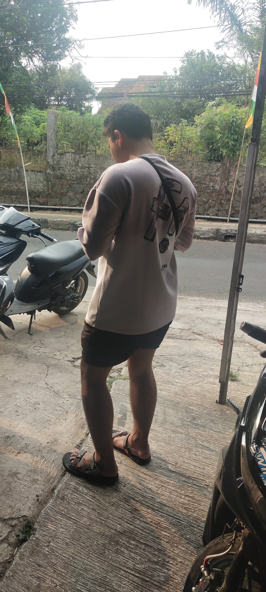 A man stands on a concrete sidewalk, facing away from the camera, with his back to a stone wall and lush green foliage. He is wearing a light-colored long-sleeved shirt with a black graphic print on the back and dark shorts. On his feet are dark-colored sandals. To his left, a black motorcycle is parked. In the background, there is a street with a small building visible and power lines overhead. The scene appears to be in an outdoor urban environment, possibly a residential area, with elements suggesting a warm, sunny day. The location is identified as Cimaung, Indonesia.