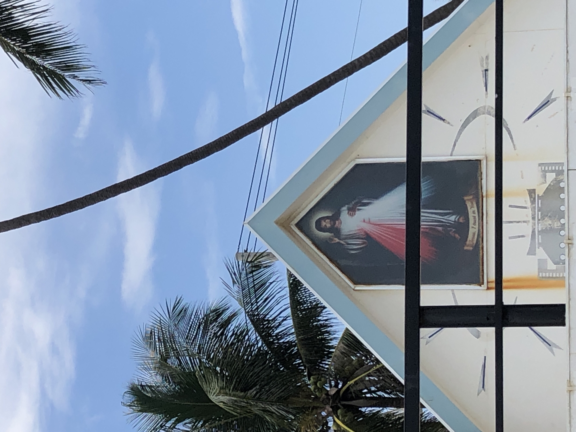 In Mũi Né, Vietnam, a religious painting of Jesus Christ, depicted with a red and white robe and the inscription "Jesus, I trust in You," is mounted on the facade of a white building with a blue trim, framed by a black metal railing. The building's triangular gable is adorned with graphic designs resembling a clock face and other abstract elements. To the left of the building, a palm tree with green coconuts is visible, and to the right, a tall palm tree trunk extends upwards, partially obscuring the bright blue sky with scattered white clouds. Overhead, dark electrical wires crisscross the frame. The overall scene suggests an outdoor setting, possibly a small chapel or shrine, under a clear sky in a tropical or subtropical location.