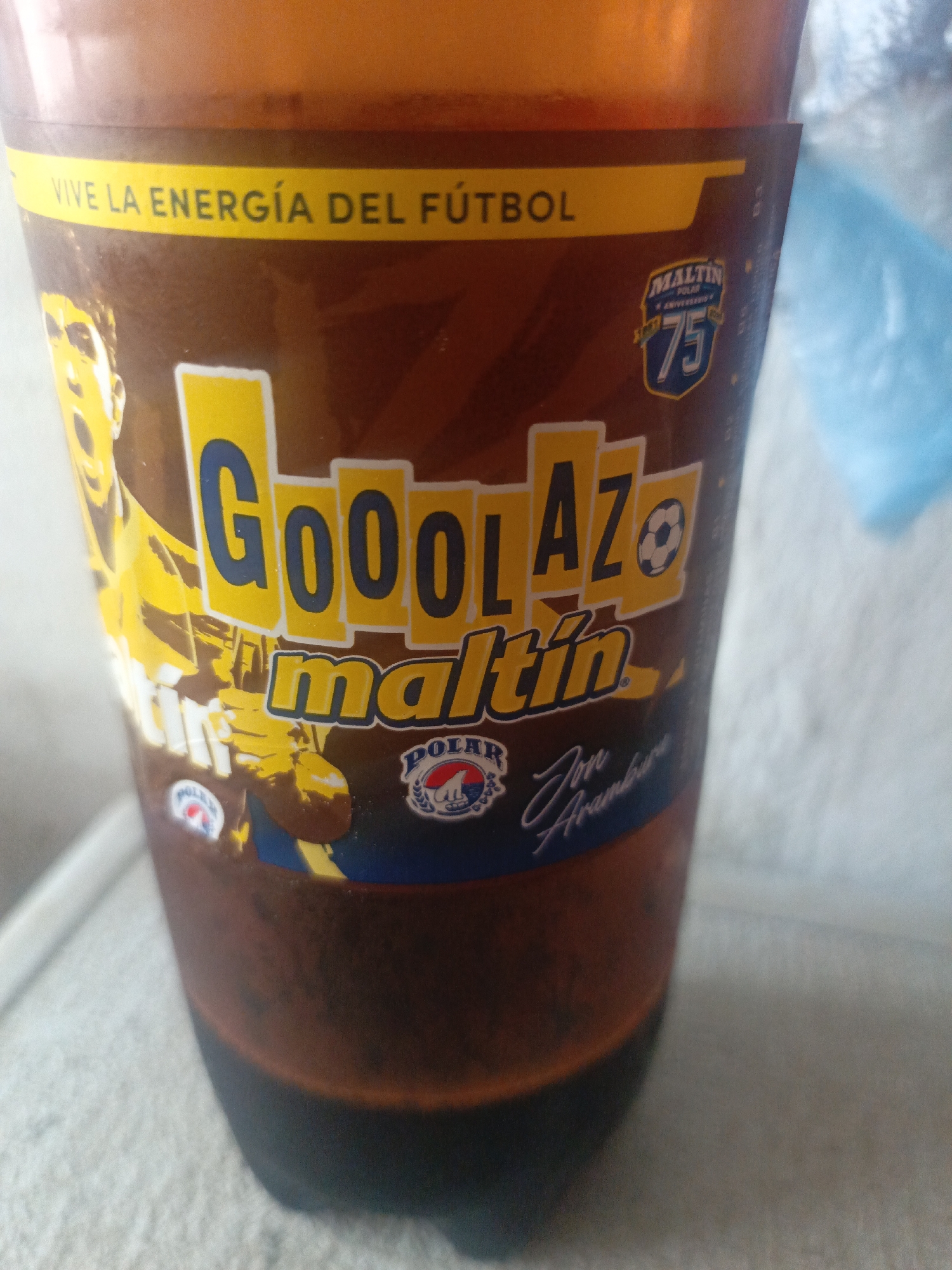 The image shows a close-up of a plastic cup filled with a dark brown liquid, likely a Maltin Polar beverage. The cup's label features the words "GooolAZo maltin" in yellow and blue lettering, along with a soccer player graphic. A slogan in Spanish, "Vive la energia del Futbol" is visible at the top. The Maltin Polar 75th anniversary logo is also present. The cup is placed on a gray surface. There is no information in the image that describes the location in the city of Juan Griego, Venezuela.