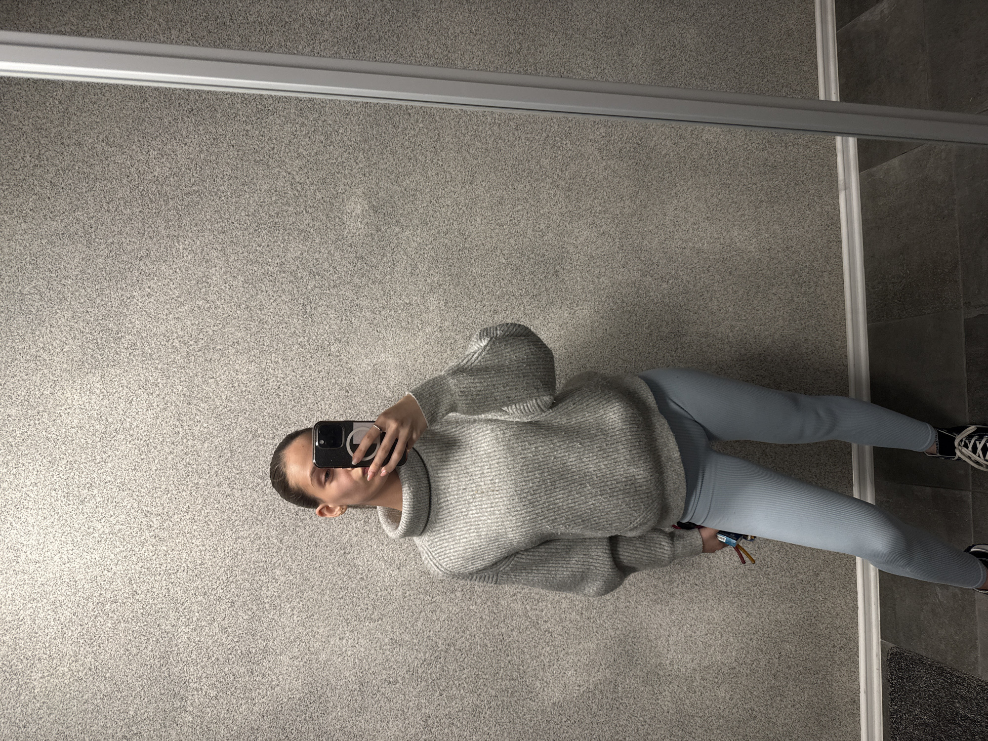 A young person, likely female, with dark hair tied back, is captured in a mirror selfie within an indoor setting. They hold a black smartphone, featuring a distinct camera array and what appears to be a lens protector ring, in their right hand, aimed towards the mirror. In their left hand, they grasp a small bundle of keys adorned with colorful tags.
The individual is dressed in casual attire: a loose-fitting, light grey, ribbed turtleneck sweater paired with light blue ribbed leggings and black sneakers with white laces. They stand on a light grey, speckled carpeted floor. To their right, there's a clear transition to a darker, tiled floor, separated by a light-colored threshold. A dark, textured mat is visible on the tiled section.
The mirror, which forms the basis of the selfie, features a thin, silver-colored track or frame running horizontally across its top edge, suggesting it might be part of a sliding door or a large, fixed wall mirror. The lighting is soft and even, typical of indoor illumination. The person maintains a neutral, focused expression, engaged in the act of taking the photo. There is no visible text within the image. The general location context provided is Sofia, Bulgaria.