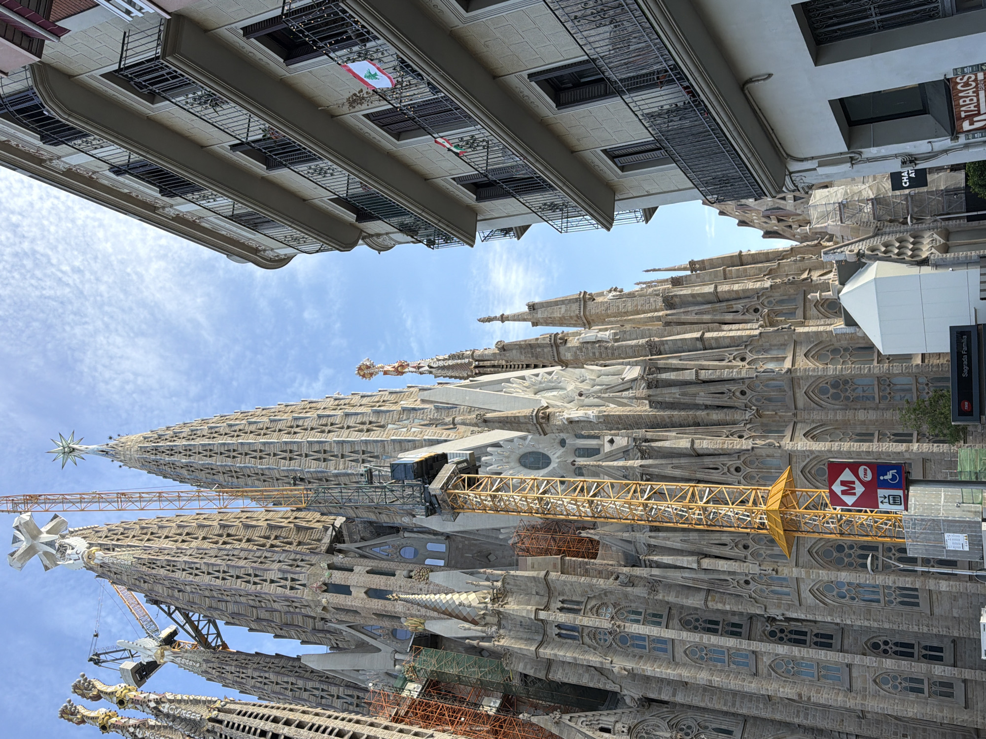 The image depicts a street view in Barcelona, Spain, with the Sagrada Familia basilica dominating the left side of the frame. The basilica, an unfinished architectural marvel, features intricate stone carvings and numerous spires, with construction cranes visible amongst its towering structures. In the foreground, a modern apartment building with balconies stands to the right of the basilica, its facade illuminated by daylight. Two Lebanese flags are visible on the balconies of this building. A yellow construction crane is prominently placed in front of the Sagrada Familia, with a TMB (Transportes Metropolitanos de Barcelona) metro sign nearby. The sky is blue with scattered clouds. The overall scene captures the juxtaposition of historical architecture, ongoing construction, and contemporary urban life in Barcelona.
