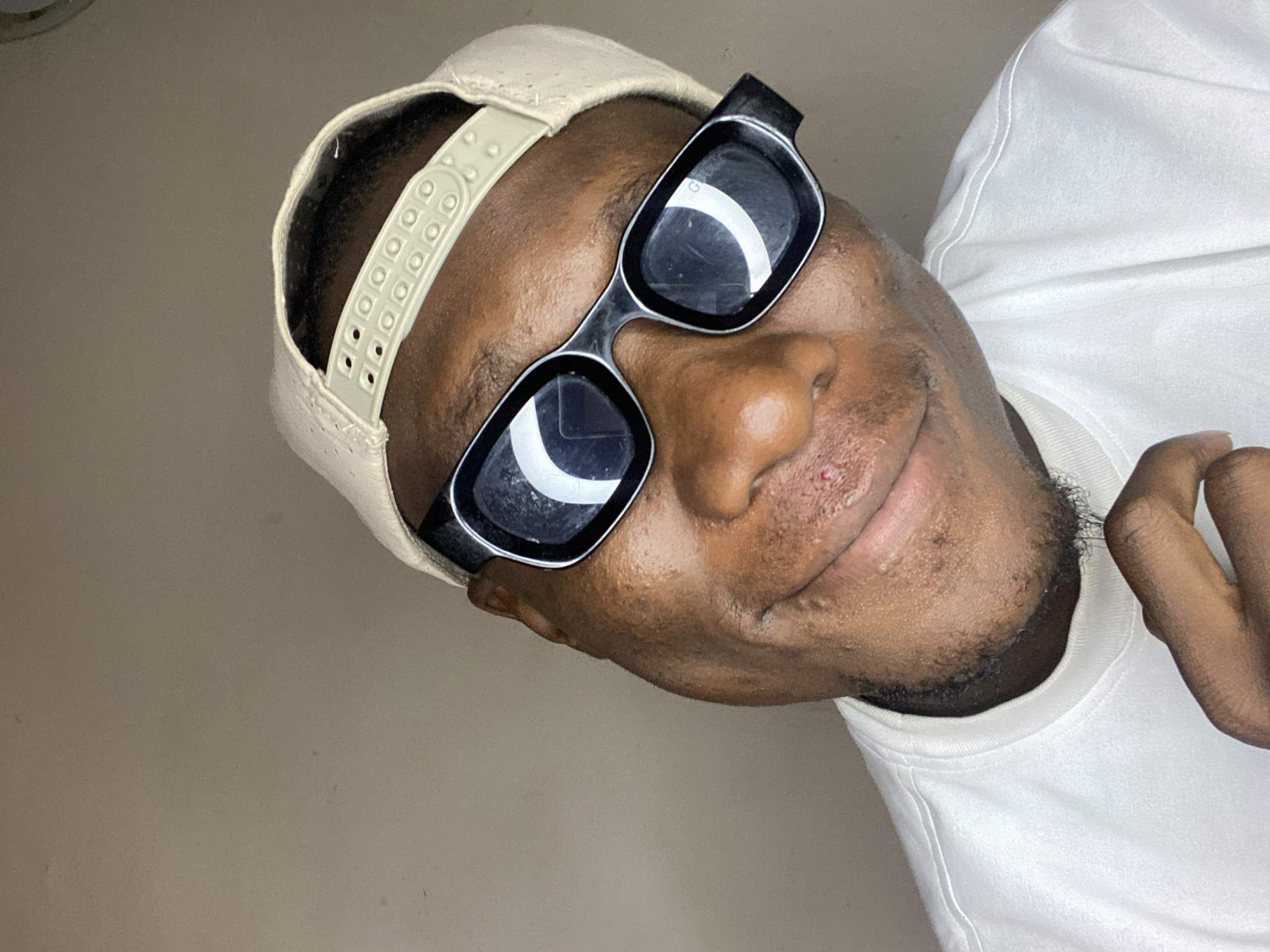 A medium close-up photograph shows a man with dark skin wearing a light-colored baseball cap and square, black-framed sunglasses. The image is angled to the left, with the man's face occupying the central part of the frame. The background is a plain, light-colored wall. The man is also wearing a white shirt or sweater. No information indicates this is in Bukuru, Nigeria.