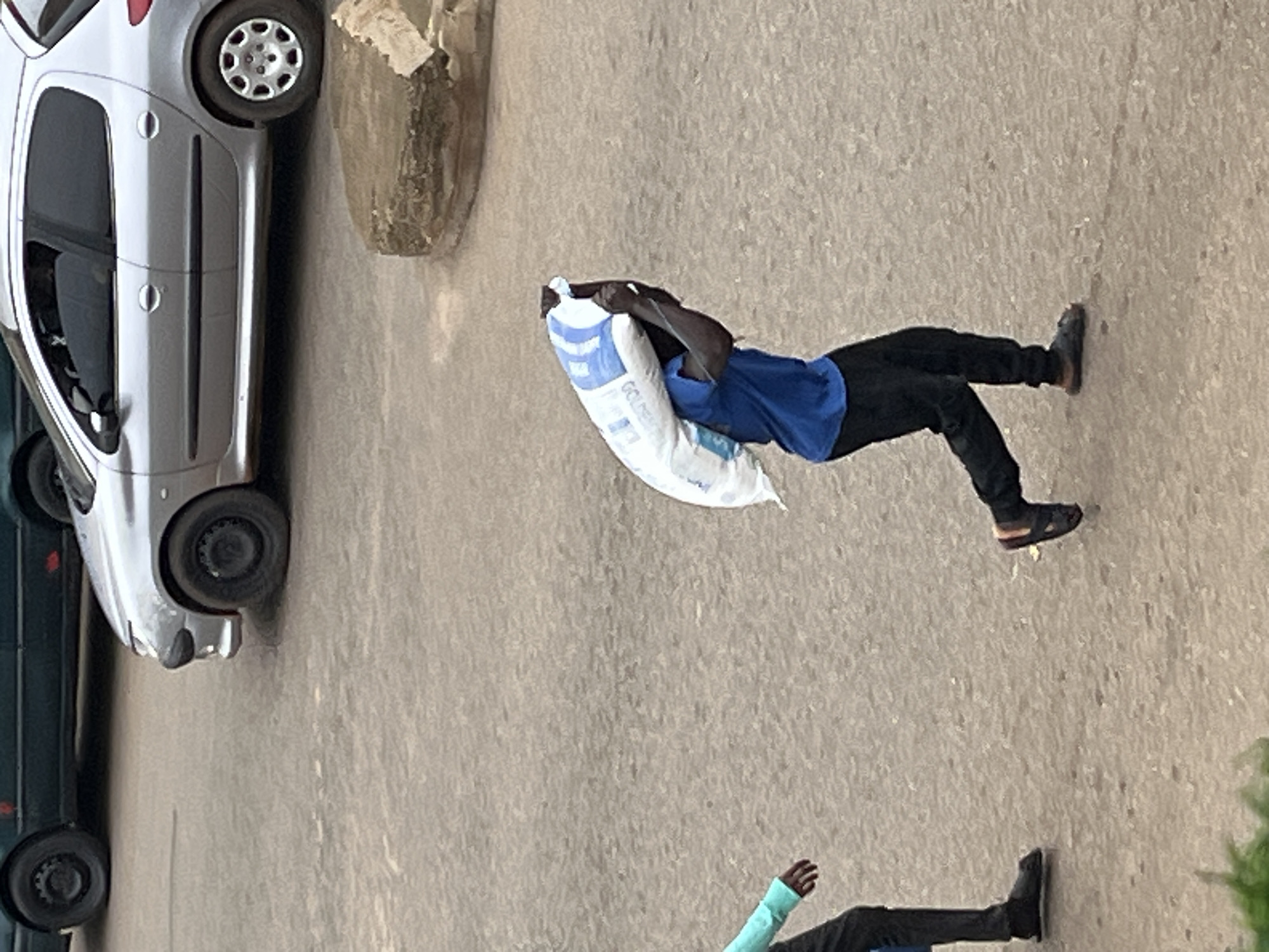 In Bukuru, Nigeria, a man in a blue t-shirt and black pants carries a large white bag filled with goods on his shoulders. He is walking on a paved surface, which appears to be a street or parking lot. To his left, another person, partially visible, wears a teal-colored shirt and dark pants, walking in the same direction. In the background, two cars are present: a silver hatchback and a dark-colored vehicle. A large, textured, light-brown rock formation is situated to the right of the silver car. The lighting suggests an overcast or diffused light condition.