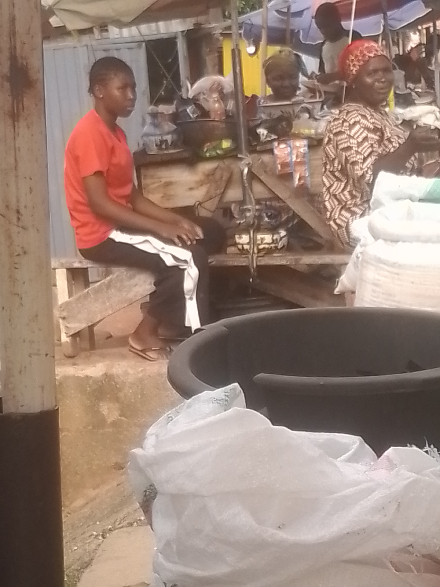 The image shows a scene at what appears to be an outdoor market or stall. In the foreground, a dark, large circular object, possibly a tub or basin, is visible. Partially obscuring the lower part of the image is a white, crinkled material, likely a bag or tarp.
The main subjects are three individuals. On the left, a young person wearing an orange t-shirt and black pants with a white stripe down the side is sitting on a wooden bench or platform. They are looking to the right. In the middle background, another person is partially visible, wearing a headscarf. To their right, an older woman wearing a patterned dress and a red headscarf is looking towards the viewer, seemingly engaged in a transaction or conversation. She is positioned behind a wooden stall.
The stall itself is made of wood and is cluttered with various items. On the stall's surface, there are some packaged goods, possibly snacks or food items, with visible red and white packaging. Behind the stall, a person is partially visible in the background, appearing to be a customer or vendor.
The setting suggests an informal marketplace. The lighting is somewhat dim, and the overall impression is that of a busy, perhaps slightly chaotic, environment. The ground appears to be concrete or dirt, with some scattered debris. The weather is not discernible, but the scene suggests daytime. There is no visible text in the image. The emotions of the subjects are difficult to ascertain, though the woman in the red headscarf appears to be smiling or speaking animatedly.