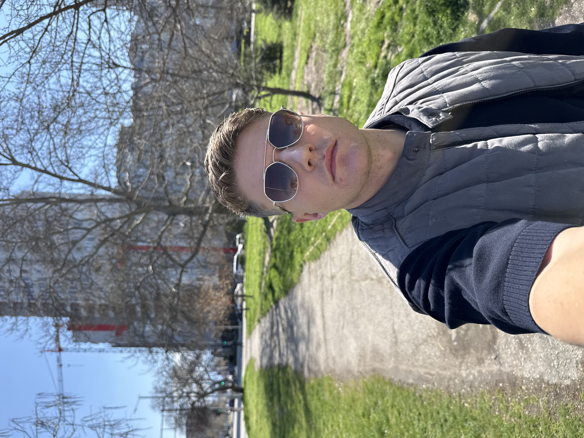 A man wearing sunglasses and a grey quilted vest over a dark blue sweater stands on a paved path in a park. He is holding the camera and taking a selfie. Behind him, there is a green grassy area, trees without leaves, and in the distance, a building under construction with a crane. The location is Plovdiv, Bulgaria. The sky is blue.