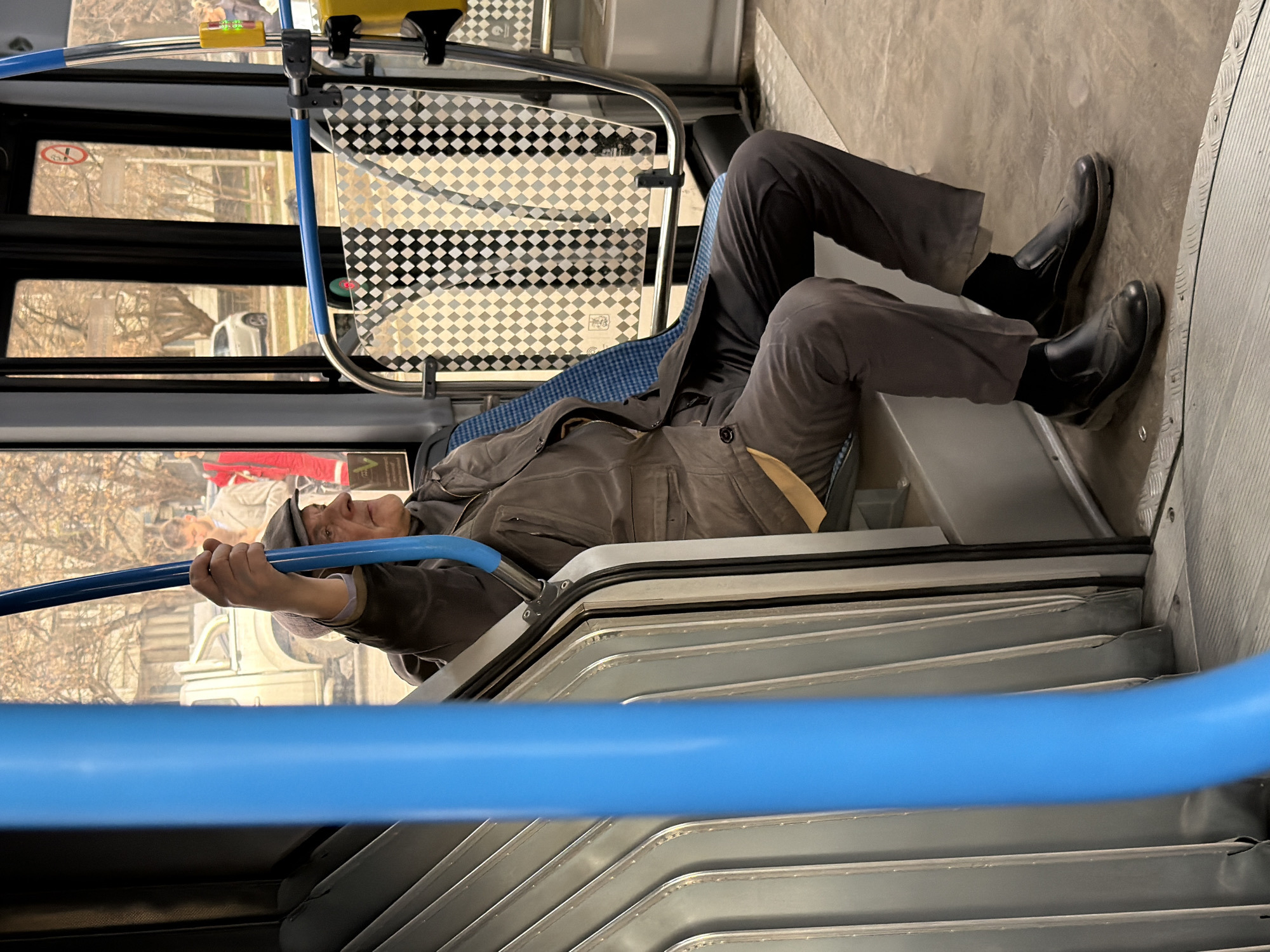 A man is seen on a bus in Khimki, Russia. The man, wearing dark trousers, black boots, a jacket and a cap, appears to be lying down across several seats. He is holding onto a blue handrail. The interior of the bus is visible, including the checkered pattern on the seatbacks, windows showing the outside scenery, and the metal seat frames.