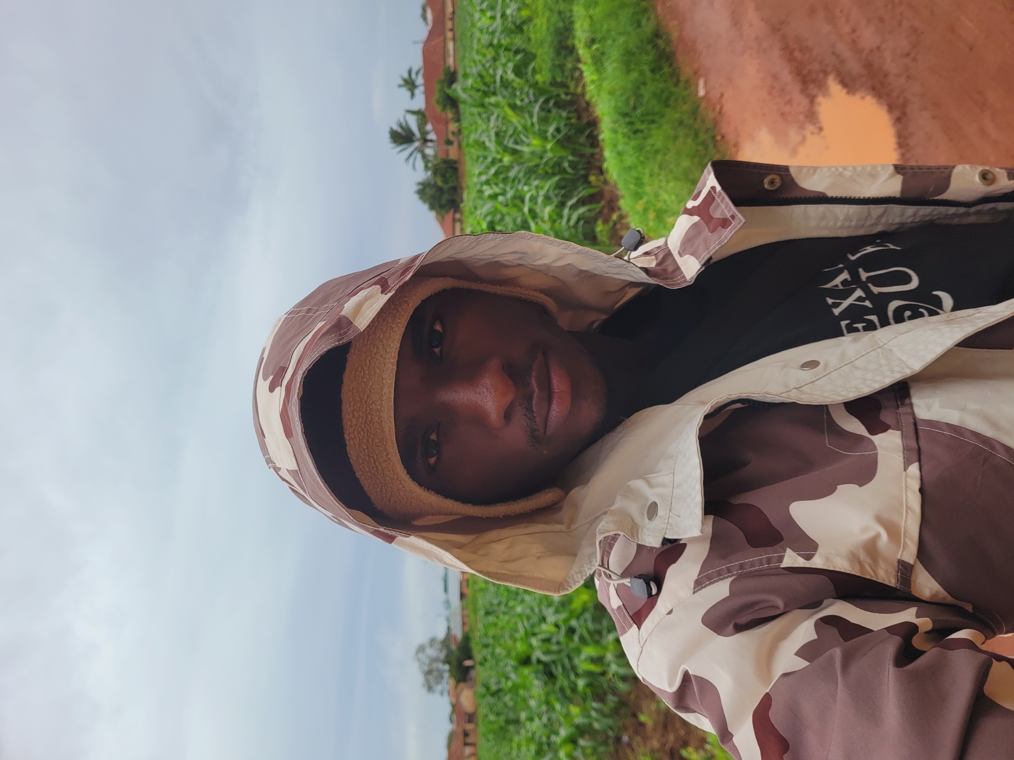 A young Black man is captured in a close-up, sideways selfie. He is wearing a camouflage jacket with a fuzzy tan lining visible around the hood. The jacket has metal snaps. Underneath the jacket, a black t-shirt with white text is partially visible. The text appears to be "EXAU". He has a serious expression.
The background suggests an outdoor, rural setting. Green crops, possibly corn, stretch into the distance, bordered by a dirt path. In the far background, there are palm trees and what appear to be buildings. The sky is overcast, indicating a cloudy or possibly rainy day. The overall lighting is soft and diffused. The image composition and the subject's attire might suggest a casual or adventurous moment. The location context places this scene in Bukuru, Nigeria.