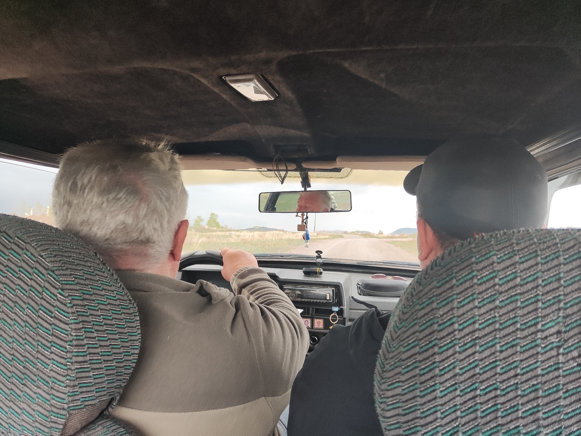 A photograph taken from the back seat of a car shows two people in the front seats. The driver on the left has short white hair and is wearing a long-sleeved beige shirt. His hands are on the steering wheel. The passenger on the right is wearing a black baseball cap and a black shirt. The car is driving on a narrow, two-lane road with a brown, grassy landscape on either side. The car's dashboard and rearview mirror are visible, along with the car's headliner. The location is Unknown, Unknown.