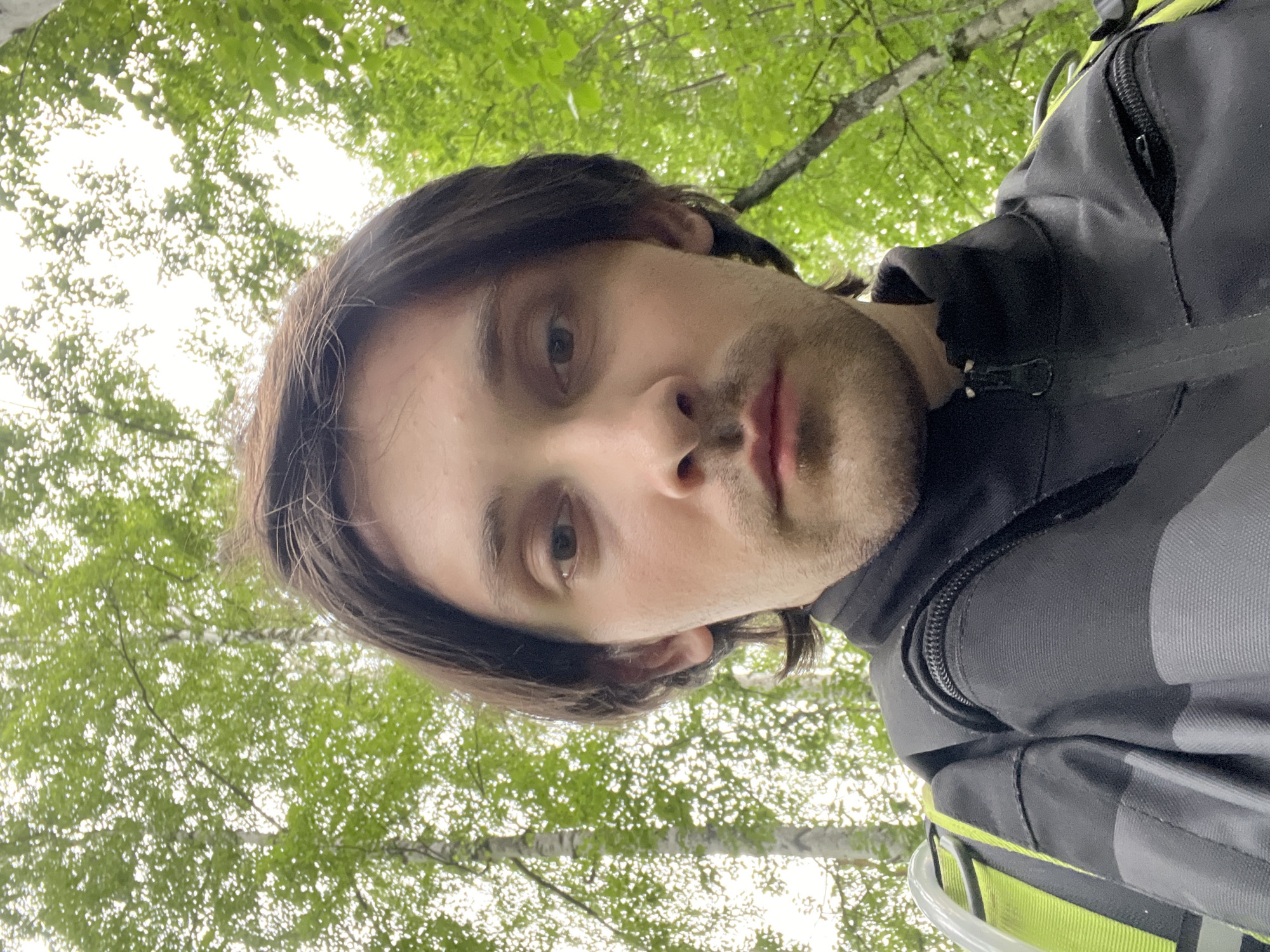 A selfie of a man with dark hair and a light beard looking upwards into a tree canopy. The background is filled with green leaves and glimpses of tree trunks, likely in a wooded area. The man is wearing a black and grey jacket with some yellow accents. No further information is available about his location beyond the trees and vegetation. No evidence indicates Vasilyevo, Russia.