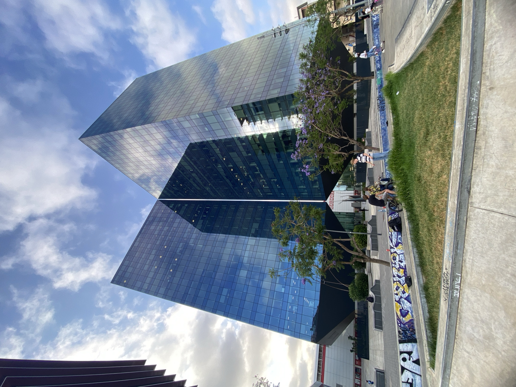 This is a low-angle, wide shot of a modern, angular glass building in Barcelona, Spain, on a partly cloudy day. The building is predominantly blue, reflecting the sky and surrounding environment. The architectural style is sharp and geometric, with large glass panels forming its exterior.
In the foreground, a section of a skatepark is visible, characterized by concrete ramps and edges. The concrete is adorned with graffiti in various colors, including blue, yellow, and purple. A patch of green grass is situated within the skatepark area.
Several individuals are present in the scene. Two people are seated on the edge of the skatepark, with one appearing to be looking at their phone. Another person is standing near a bicycle, which is leaning against the graffiti-covered wall. A person in a striped shirt and jeans is walking, and another person is on a skateboard. Further in the background, more people are visible, including one walking near the building.
Trees with purple flowers are planted around the skatepark area, adding a touch of nature to the urban landscape. The building's reflection is prominent on its glass facade, mirroring the sky, clouds, and parts of the surrounding architecture. The overall impression is of a vibrant, contemporary urban space that blends modern architecture with recreational activities and public art.